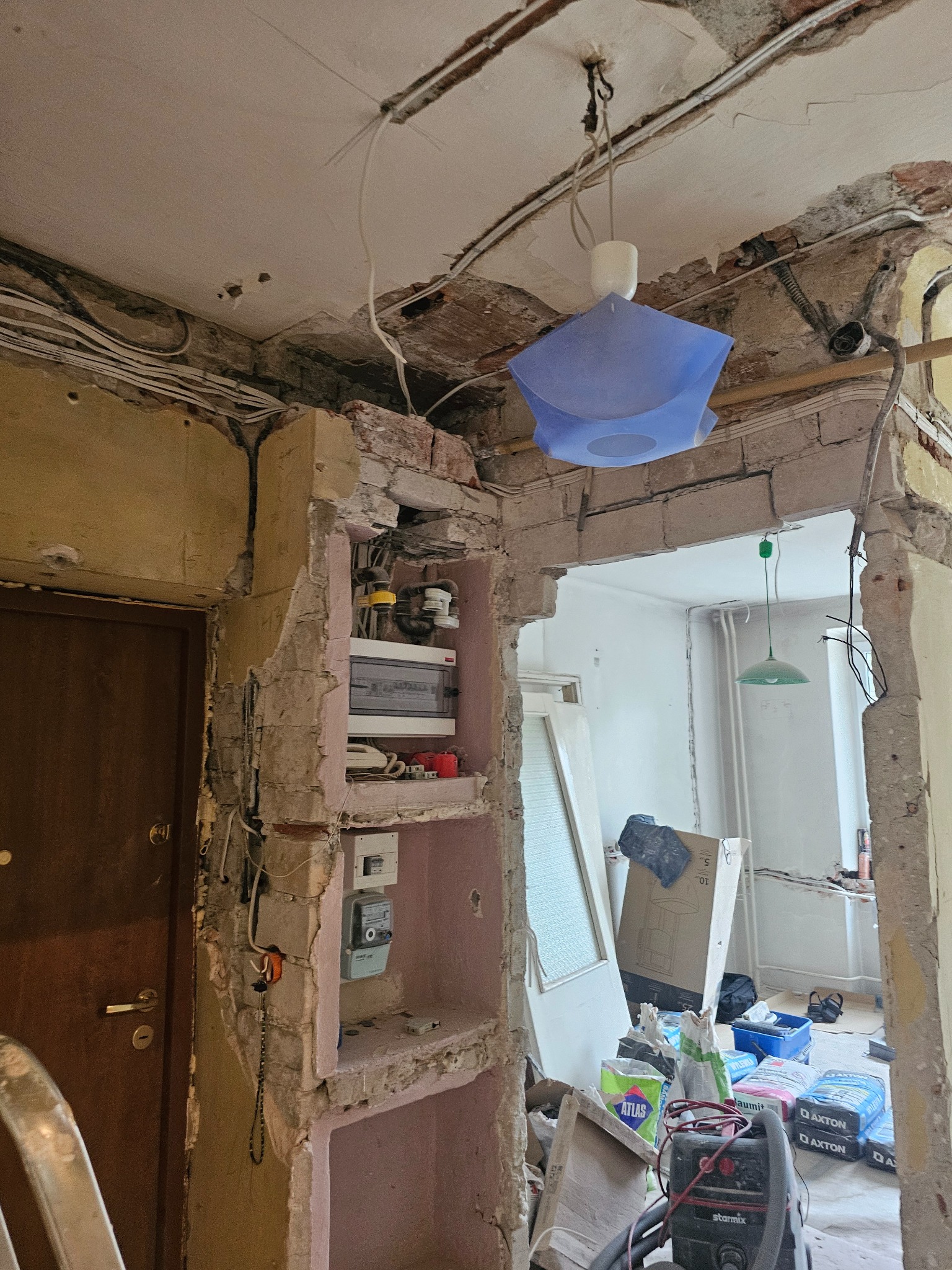 Demontaż ścian w mieszkaniu. Widoczne kable, cegły, prowizoryczne oświetlenie i materiały budowlane. Fragmentaryczny widok na zdemolowane wnętrze w trakcie prac.