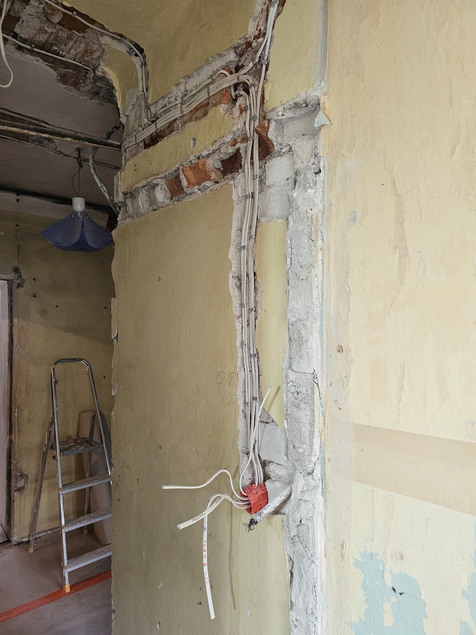 Ściana w trakcie remontu: odsłonięta cegła, instalacja elektryczna w peszlach, fragment ściany z resztkami starej farby. Widoczna drabina i wisząca lampa.