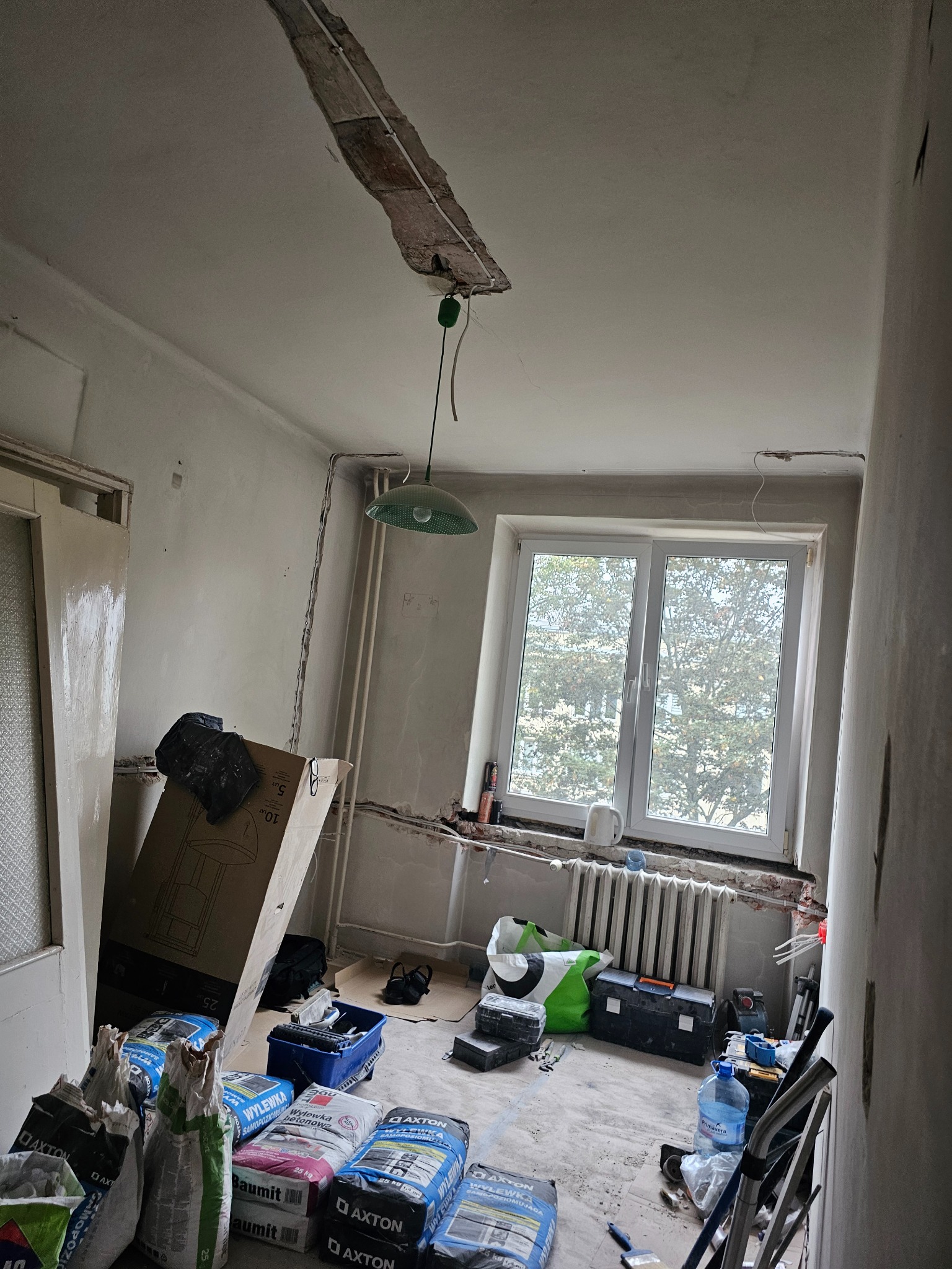 Wnętrze w trakcie remontu: widoczne worki z zaprawą, narzędzia, odsłonięty fragment sufitu i grzejnik. Przygotowania do prac wykończeniowych w pomieszczeniu.