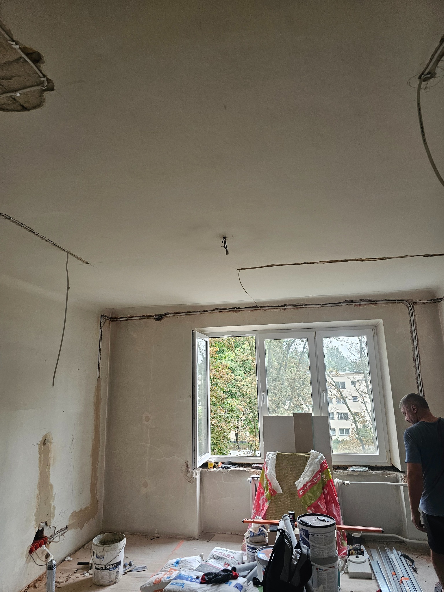 Pokój w trakcie remontu: ściany przygotowane do malowania, widoczne przewody elektryczne, okno z widokiem na zieleń, narzędzia i materiały budowlane.