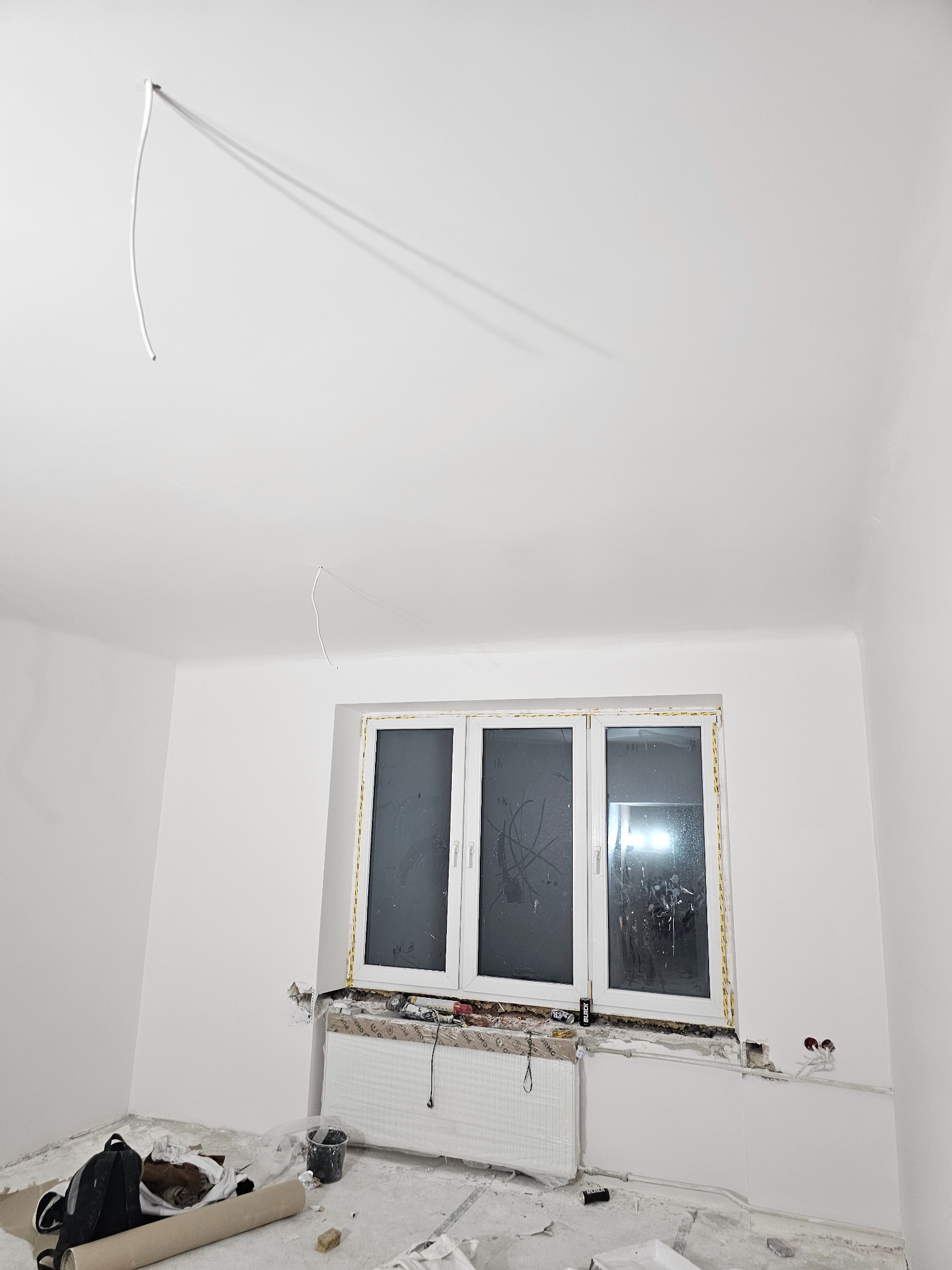 Pokój w trakcie prac wykończeniowych: świeżo pomalowane ściany na biało, okno zabezpieczone folią, widoczne narzędzia i materiały malarskie, wystający kabel z sufitu.