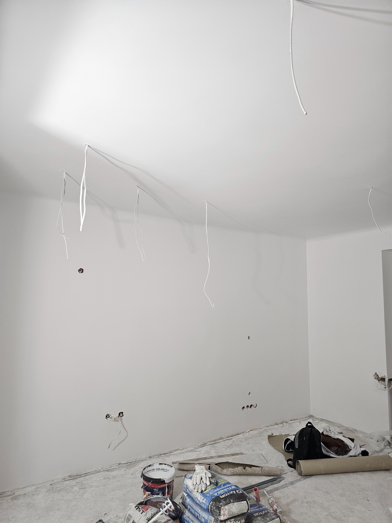 Białe ściany po szpachlowaniu, widoczne kable zwisające z sufitu, worki z gładzią i narzędzia malarskie na podłodze. Jasne wnętrze w trakcie prac wykończeniowych.