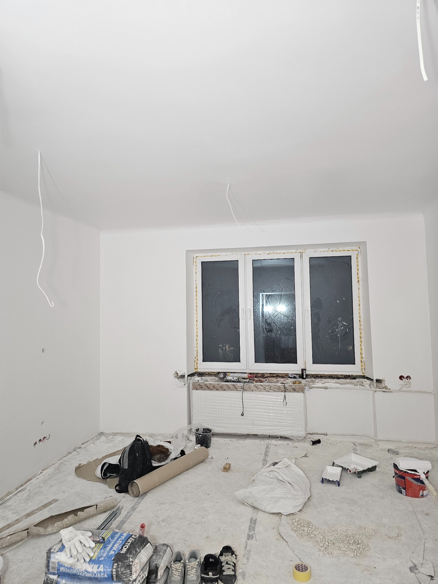 Wnętrze po malowaniu na biało. Widoczne zaklejone okno, grzejnik owinięty folią, narzędzia malarskie i worki z zaprawą na podłodze. Kabel zwisa z sufitu.