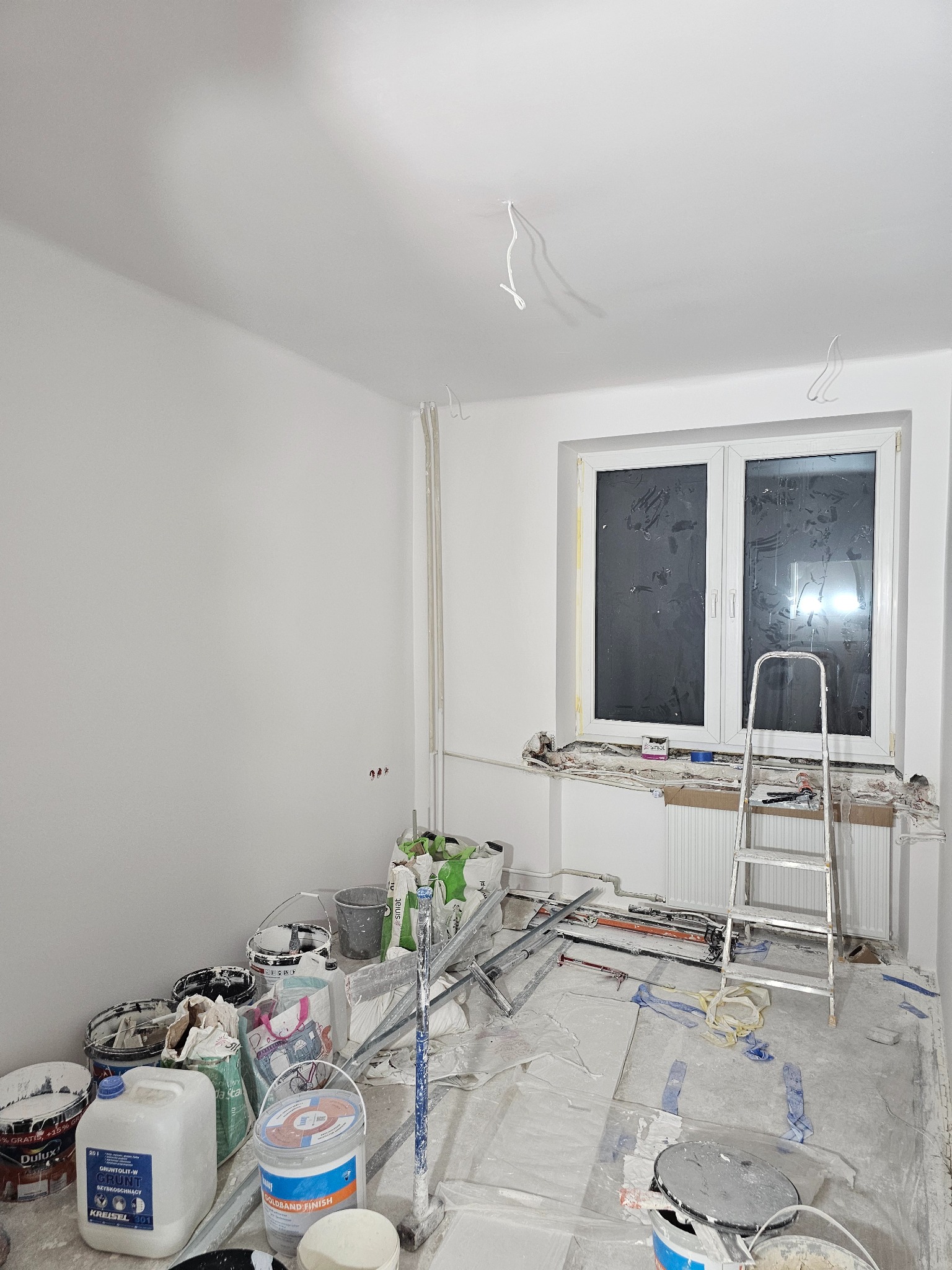 Pokój po malowaniu, z widocznymi śladami prac: zakryte okno, drabina, farby. Świeżo pomalowane ściany na biało. Etap wykończeniowy przed sprzątaniem.