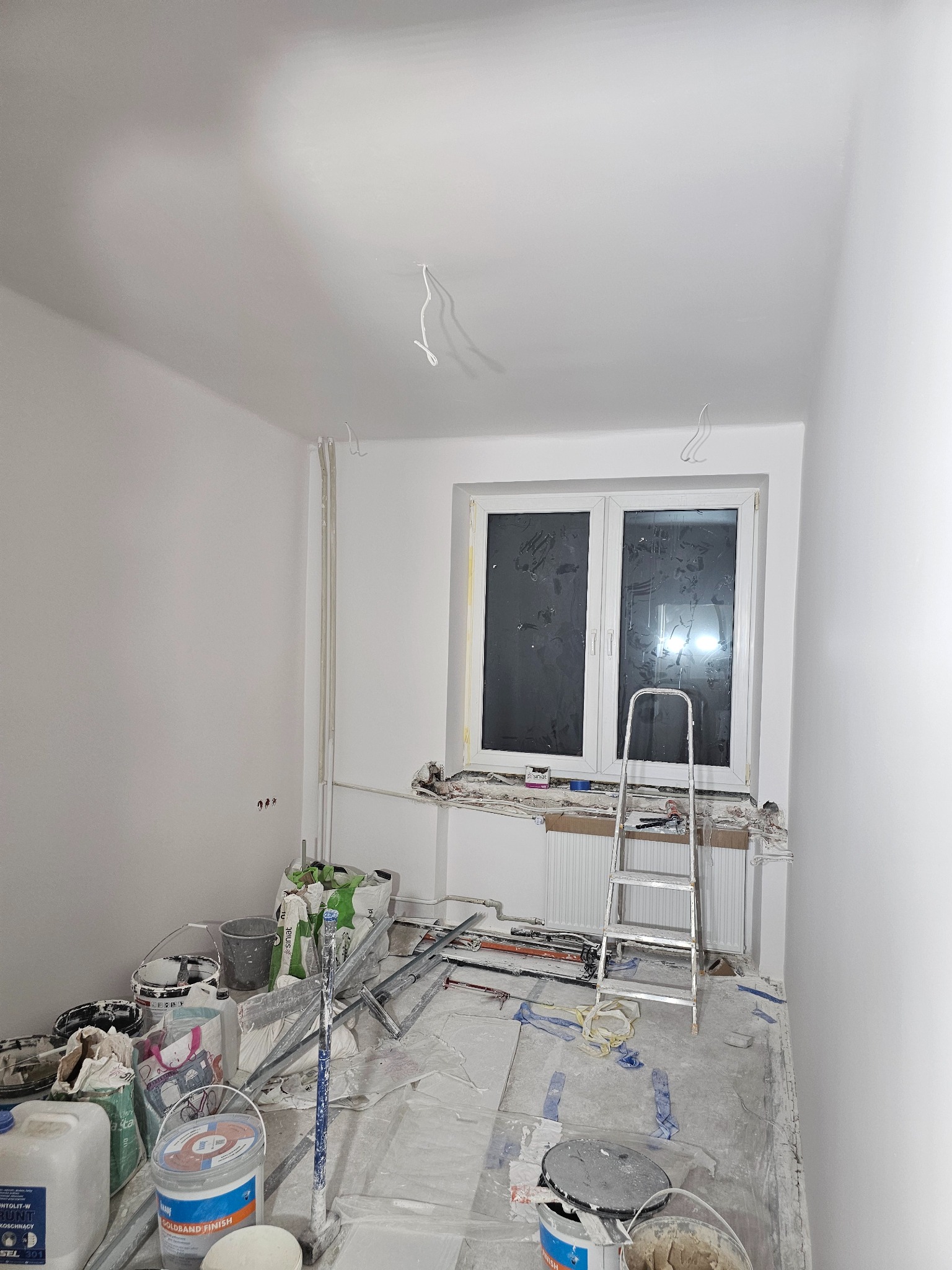 Pokój po szpachlowaniu i malowaniu, widoczne narzędzia, drabina i okno z folią ochronną. Ściany i sufit świeżo pomalowane na biało. Podłoga zabezpieczona folią.