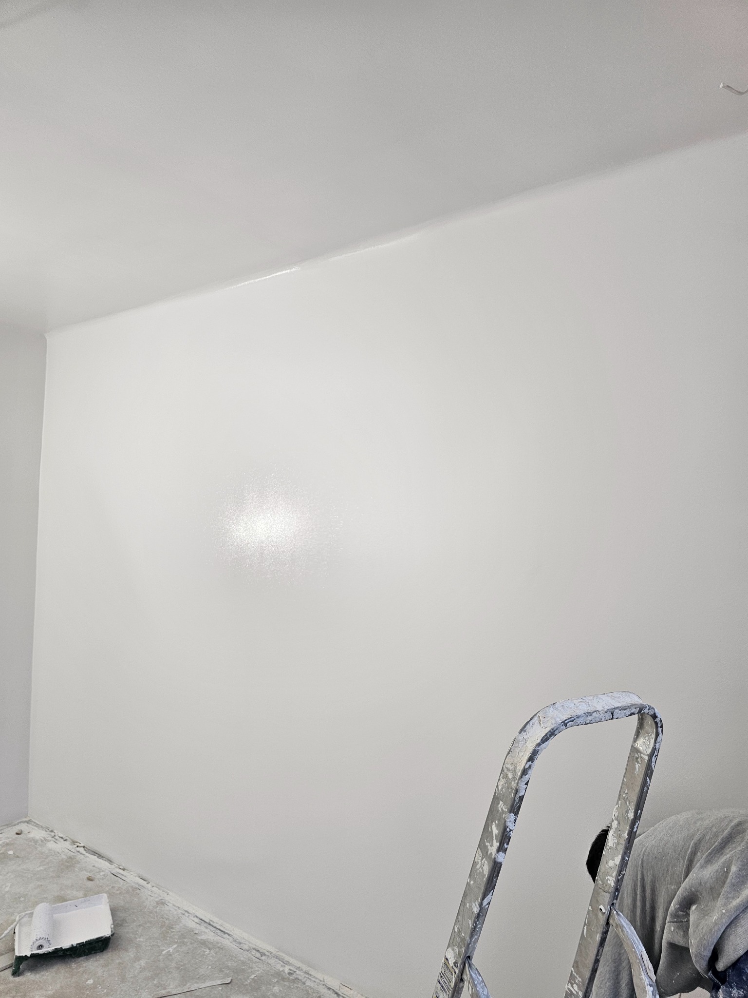 Ściana pomalowana na biało z widoczną drabiną malarską i wałkiem na podłodze. Świeżo pomalowane wnętrze z połyskiem na ścianie w centralnej części kadru.