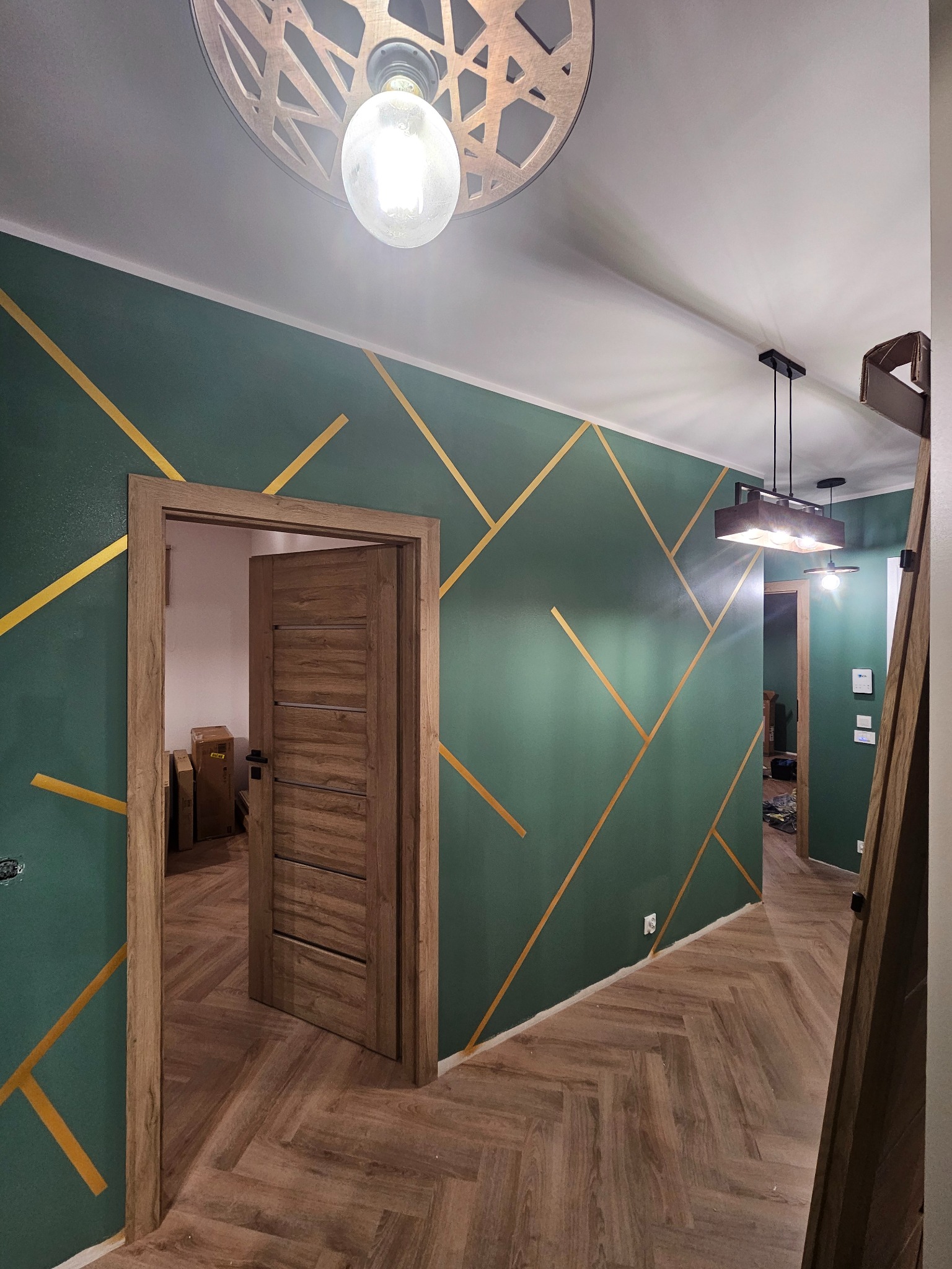 Korytarz z zielonymi ścianami i złotymi pasami, drewniana podłoga w jodełkę, widoczne drzwi i lampy sufitowe. Dekoracyjne wykończenie ścian.
