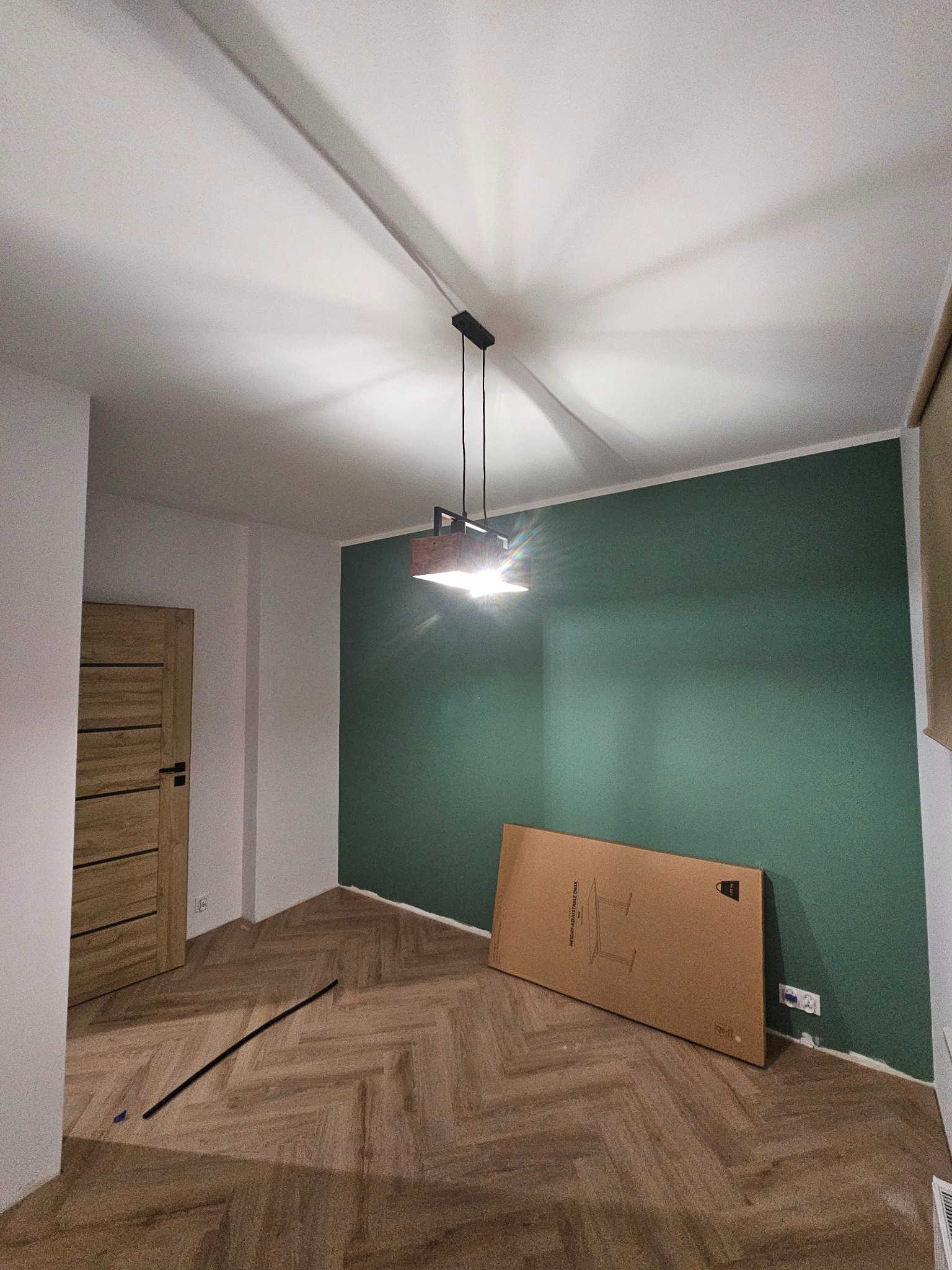 Pokój po malowaniu ścian na zielono, z parkietem w jodełkę. Widoczna lampa sufitowa, kartonowe opakowanie i listwa przypodłogowa. Świeżo wykończone wnętrze.