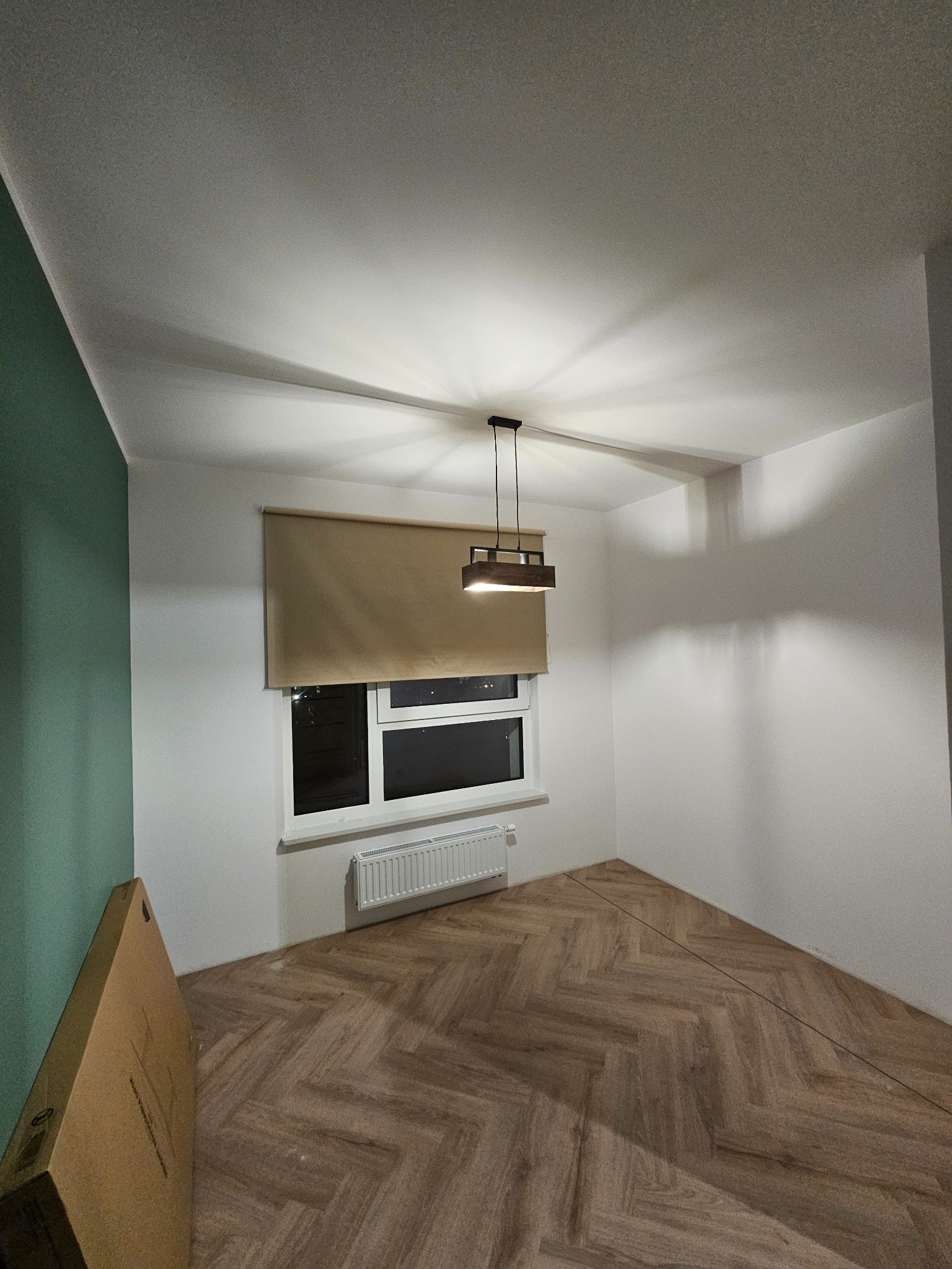 Wnętrze po remoncie: parkiet w jodełkę, białe ściany z akcentem zieleni, nowoczesna lampa. Widok na okno z zasłoniętą roletą i grzejnik.