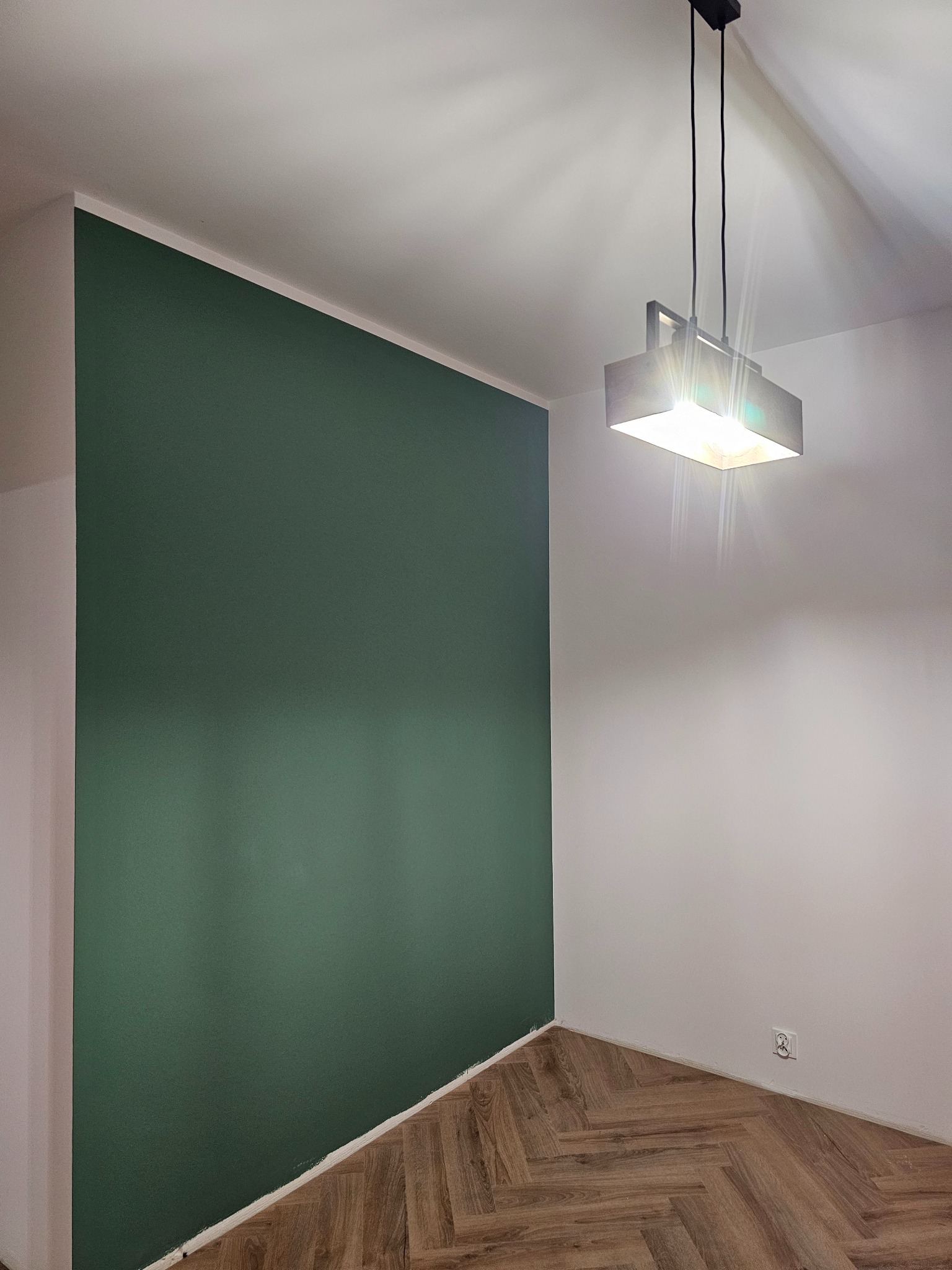 Pomieszczenie po malowaniu: ściana w odcieniu butelkowej zieleni kontrastuje z białą ścianą i parkietem w jodełkę. Nowoczesna lampa sufitowa.