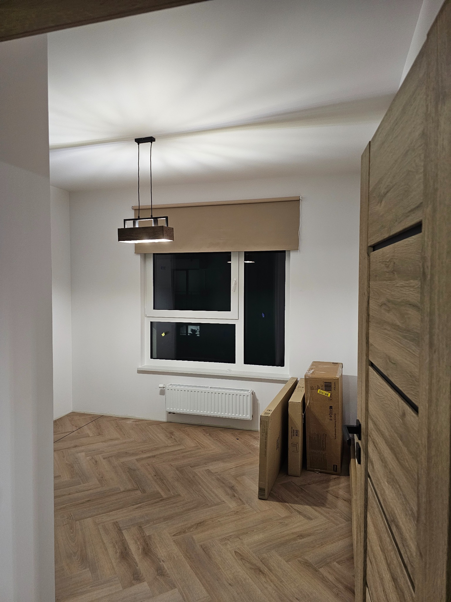 Pokój po remoncie: parkiet w jodełkę, białe ściany, okno z roletą, nowoczesna lampa. Częściowo widoczne drzwi i kartony z wyposażeniem.