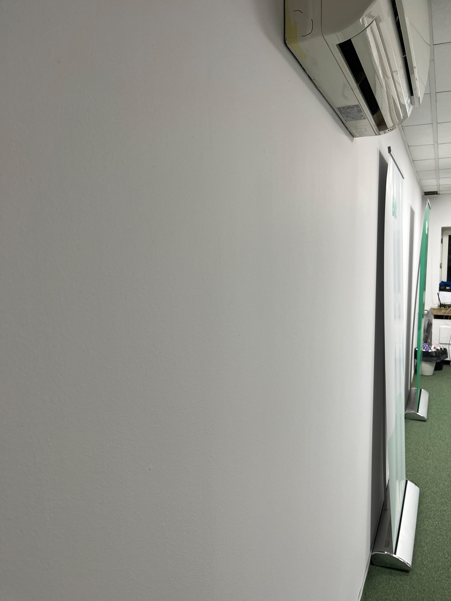 Gładka, świeżo pomalowana ściana w biurze, z klimatyzatorem pod sufitem i roll-upami w tle. Minimalistyczne wnętrze po malowaniu, jasne oświetlenie.