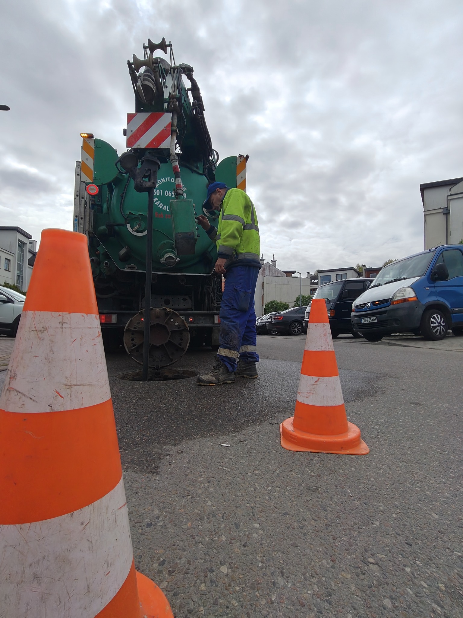 Pracownik w kombinezonie przy zielonym wozie asenizacyjnym, zabezpieczonym pomarańczowymi pachołkami, podczas czyszczenia studzienki kanalizacyjnej na ulicy.