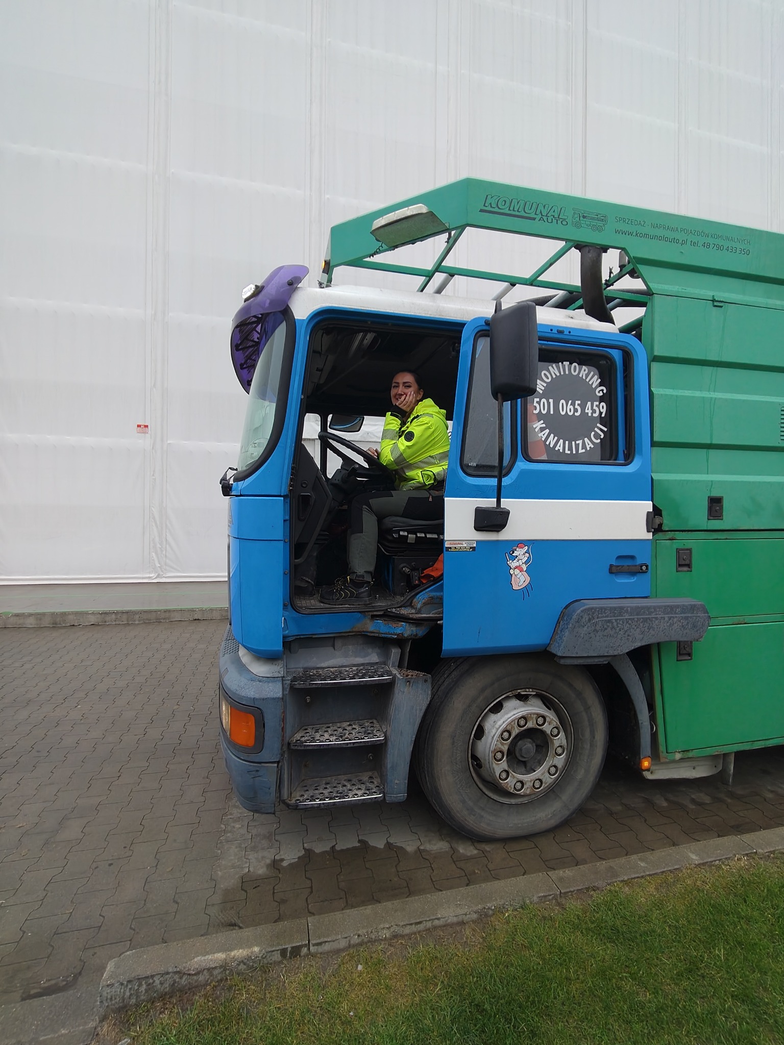 Kierowca w kabinie niebiesko-zielonego pojazdu do monitoringu kanalizacji z numerem telefonu na szybie, zaparkowanego na brukowanym placu.