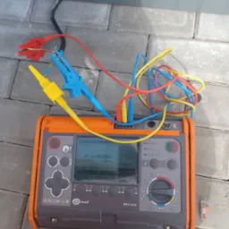 Miernik MPI-530 marki Sonel na kostce brukowej, z podłączonymi przewodami pomiarowymi zakończonymi krokodylkami. Widoczne parametry pomiarowe na ekranie.