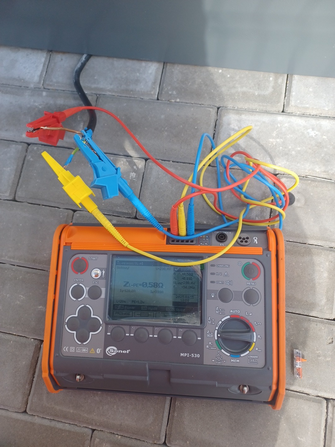 Miernik MPI-530 marki Sonel na kostce brukowej, z podłączonymi przewodami pomiarowymi zakończonymi krokodylkami. Widoczne parametry pomiarowe na ekranie.