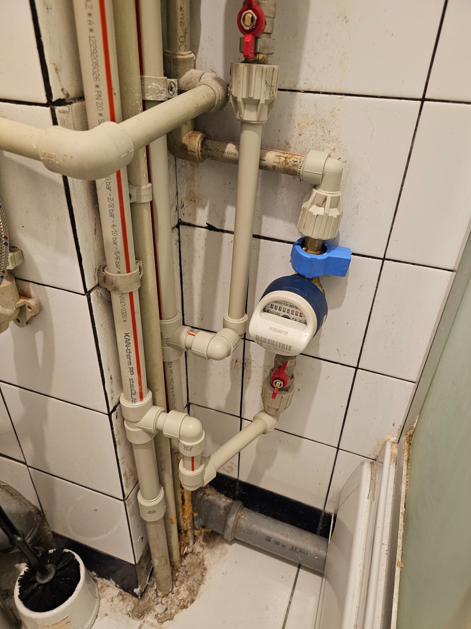 Instalacja wodna z licznikiem w łazience z białymi kafelkami. Widoczne rury z tworzywa sztucznego i metalu, zawory z czerwonymi pokrętłami i niebieski zawór.