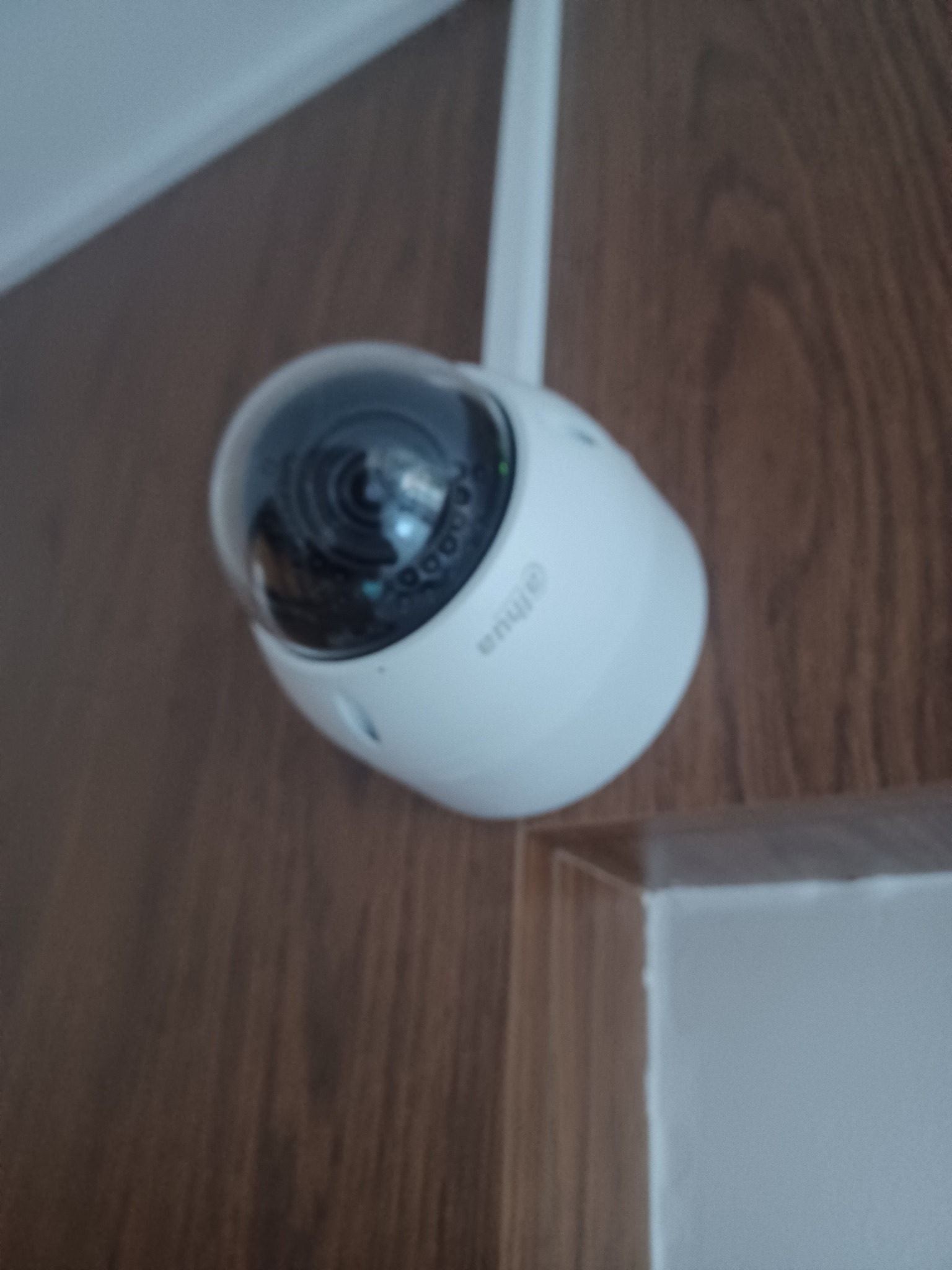 Kamera IP Dahua z anteną na tle drewnianej ściany i białego narożnika. Widoczny obiektyw kamery i logo producenta. Kamera skierowana w dół.