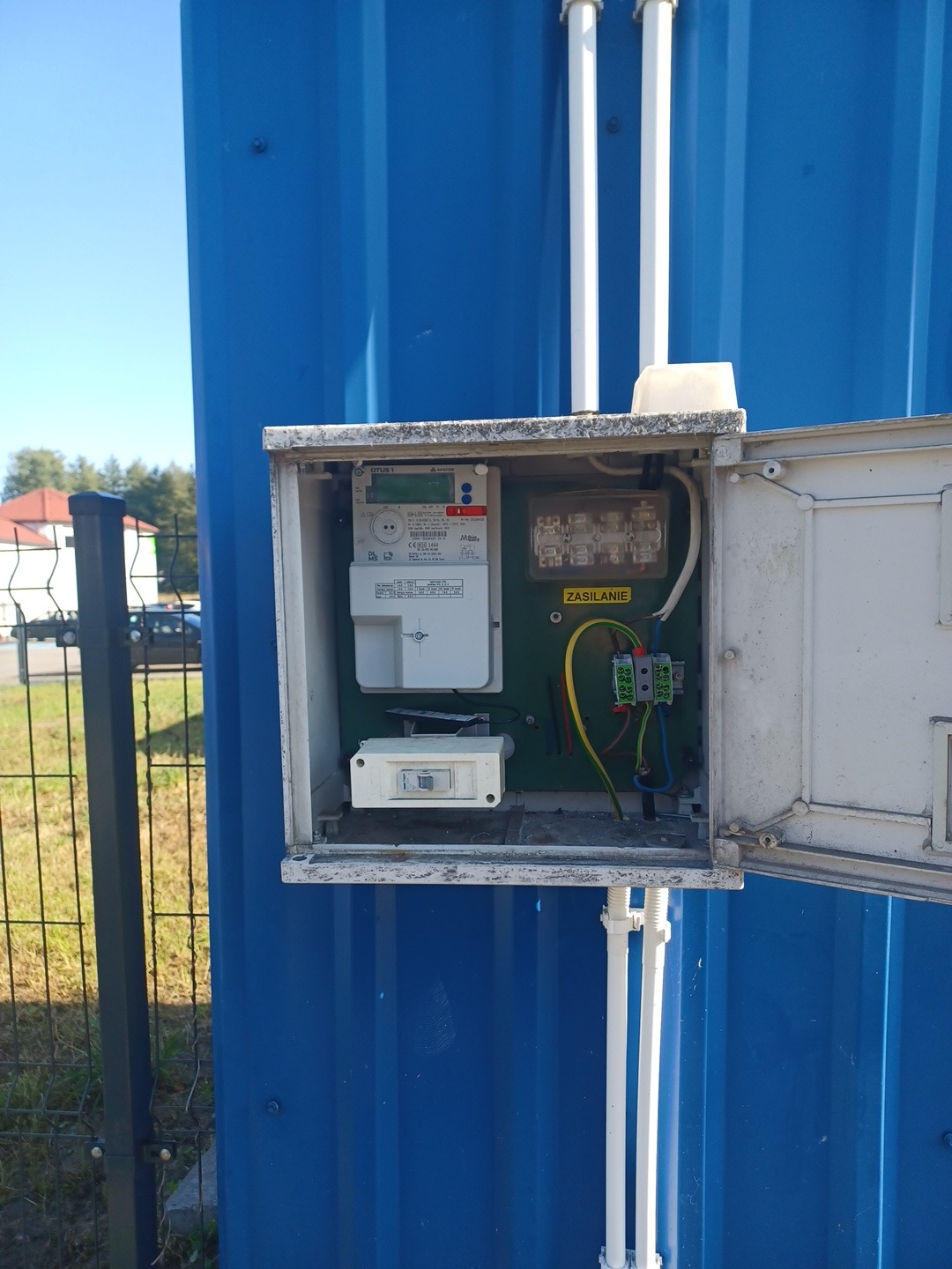 Otwarta skrzynka elektryczna z licznikiem na niebieskiej blasze, widoczne przewody, bezpieczniki i elementy instalacji. Na zewnątrz rury i ogrodzenie.