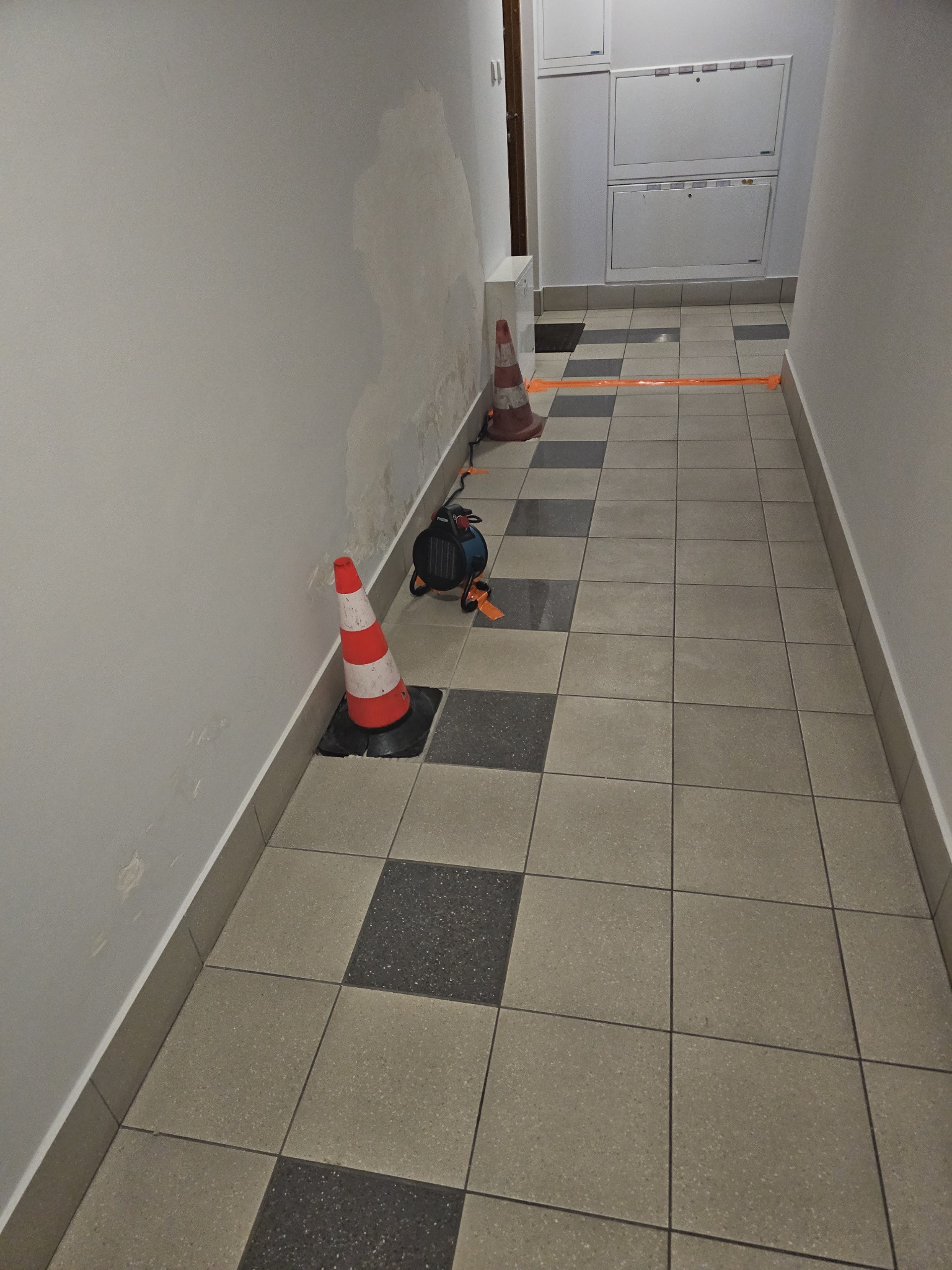 Korytarz w trakcie malowania: zabezpieczona podłoga taśmą, dwa pachołki, nagrzewnica. Ślady wilgoci na ścianie wskazują na wcześniejsze prace.