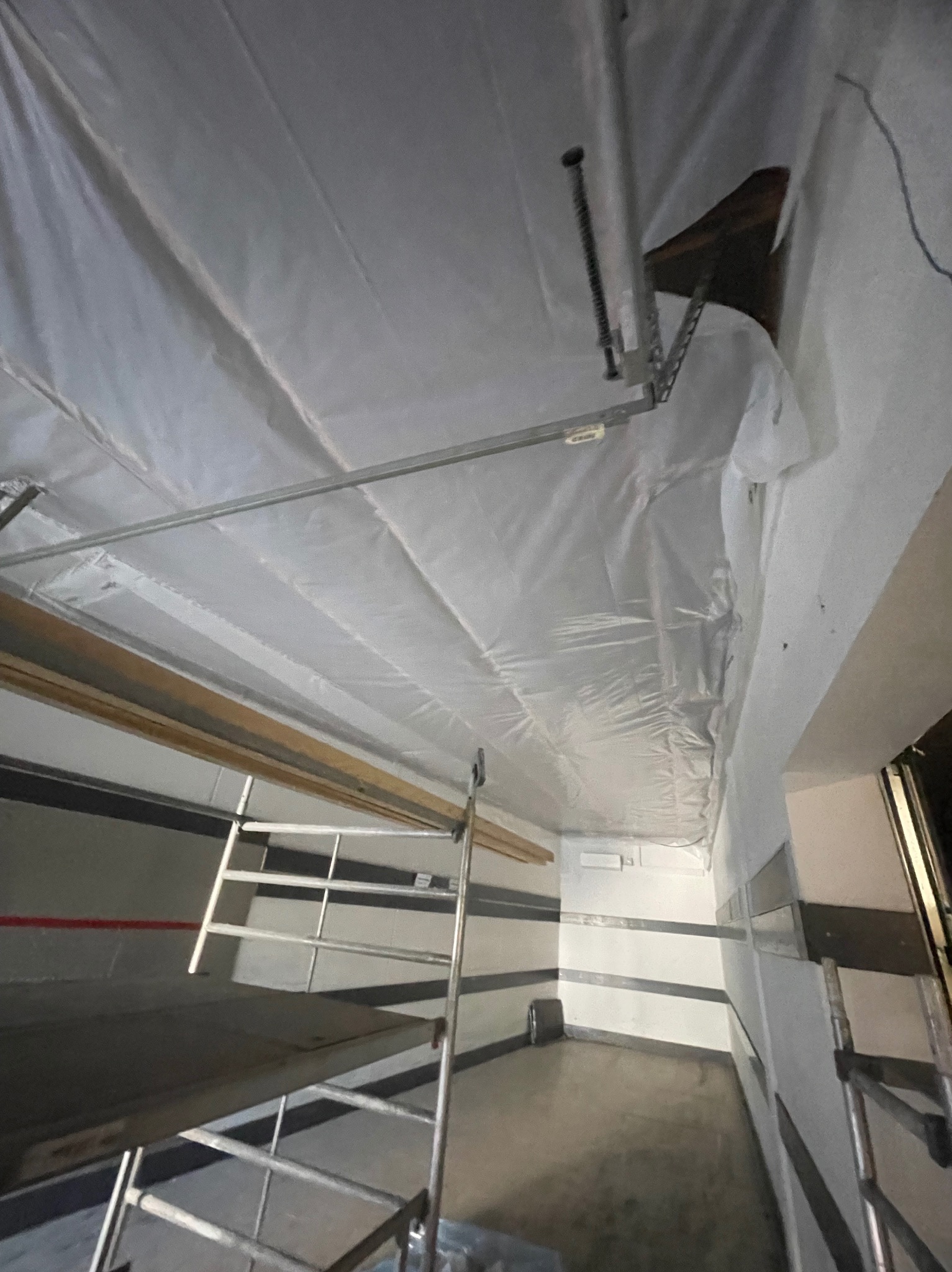 Przygotowanie do prac wykończeniowych: folia ochronna na suficie, drabina i rusztowanie w pomieszczeniu z poziomymi pasami na ścianach.