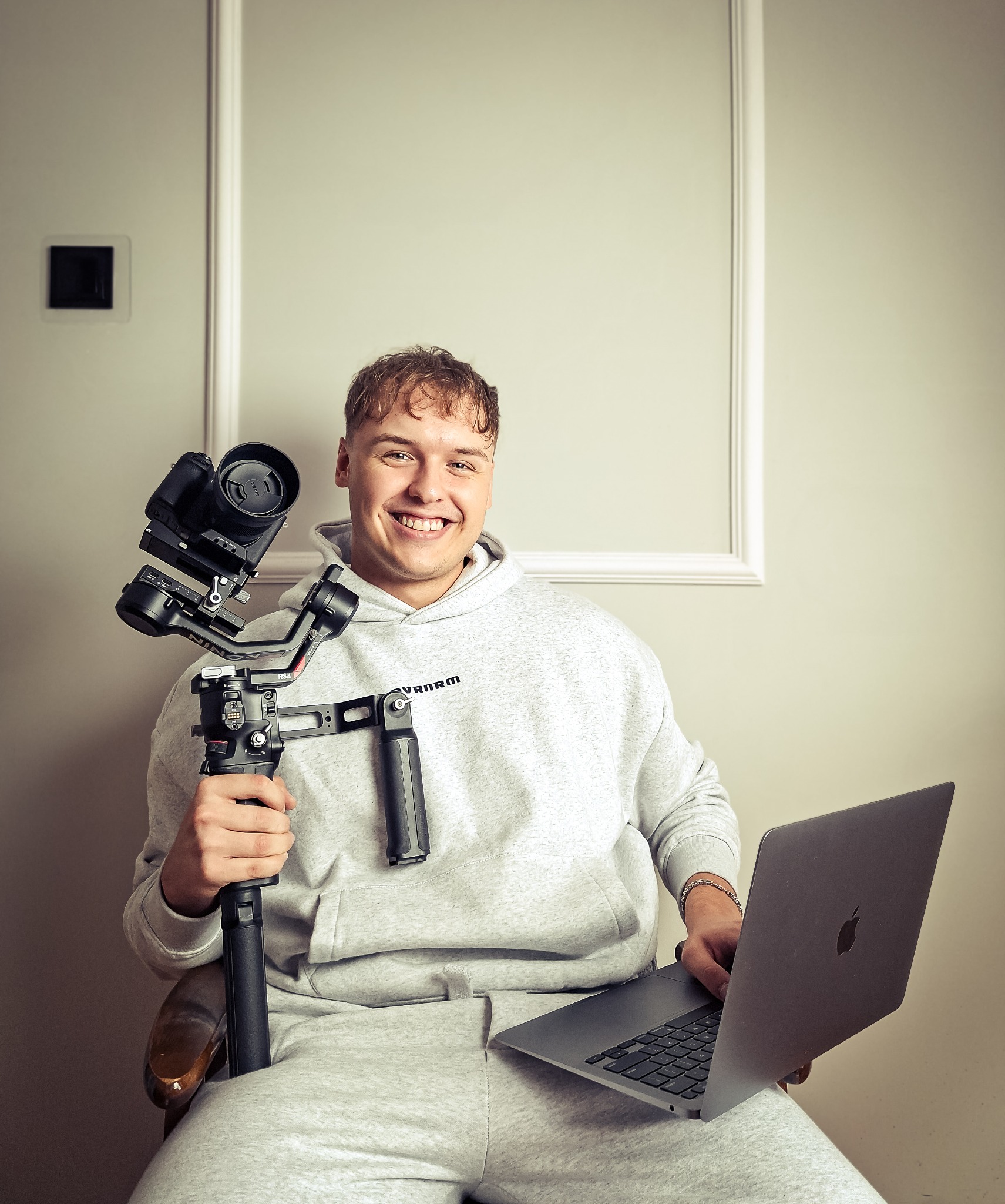 Uśmiechnięty kamerzysta w dresie siedzi z laptopem i stabilizatorem DJI RS 4 z kamerą, prezentując narzędzia pracy w jasnym wnętrzu.