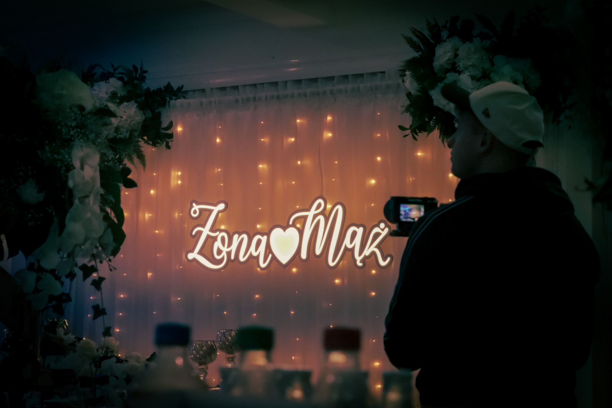 Kamerzysta filmuje neonowy napis 'Żona Mąż' z sercem, dekoracje kwiatowe i girlandy na tle. Widoczne szkło i butelki na pierwszym planie, ciemne otoczenie.