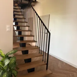 Nowoczesne schody metalowe z drewnianymi stopniami i czarną balustradą. Parkiet w jodełkę i roślina doniczkowa w tle. Minimalistyczny design.