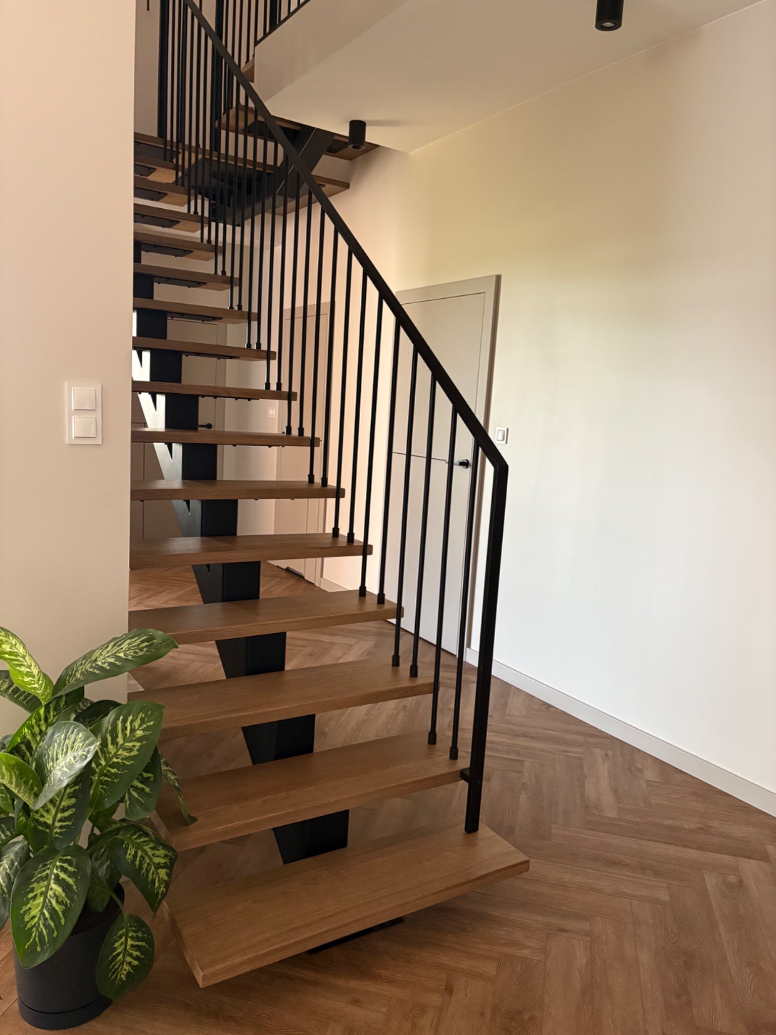 Nowoczesne schody metalowe z drewnianymi stopniami i czarną balustradą. Parkiet w jodełkę i roślina doniczkowa w tle. Minimalistyczny design.
