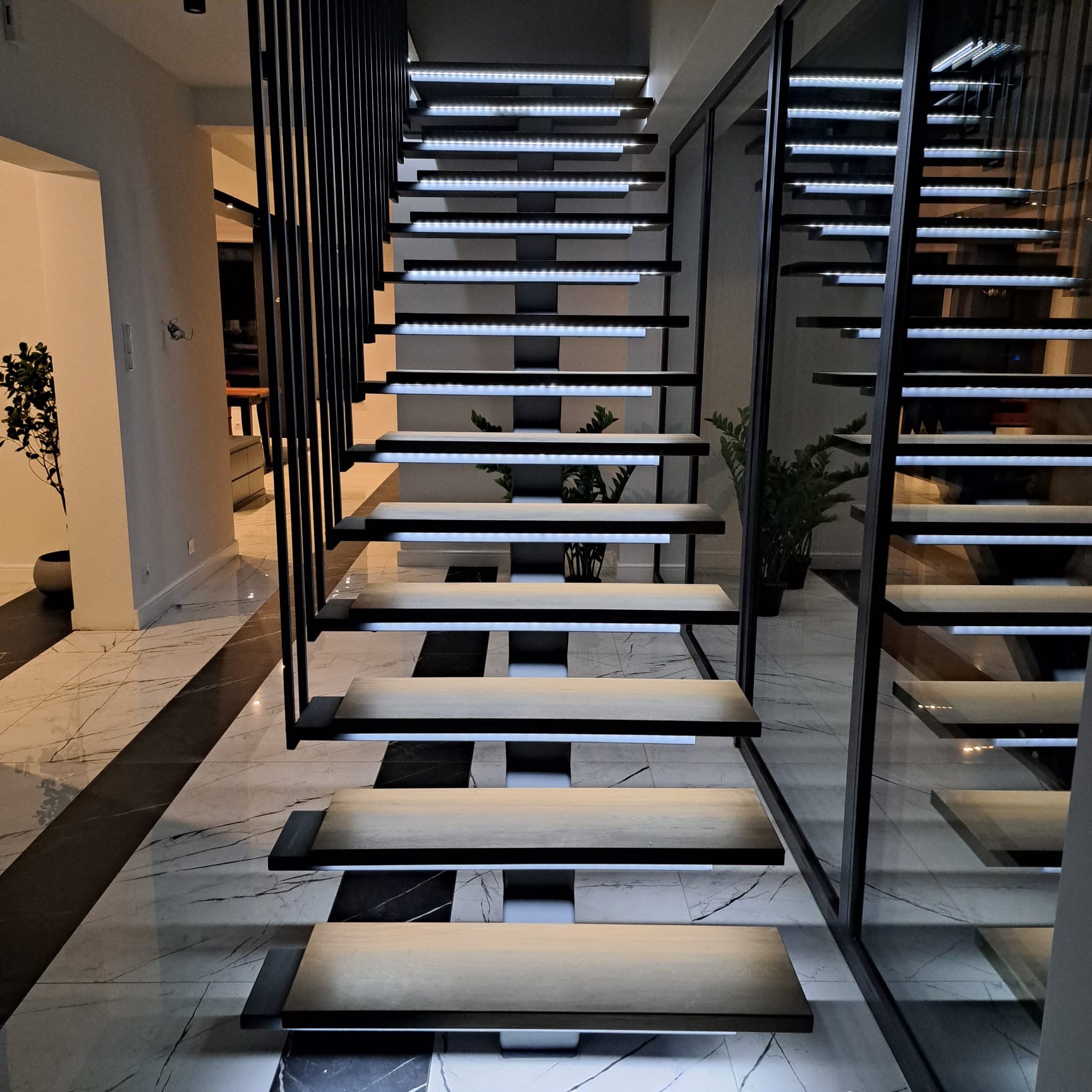 Nowoczesne, podświetlane schody z metalowymi barierkami i drewnianymi stopniami na marmurowej posadzce w minimalistycznym wnętrzu.