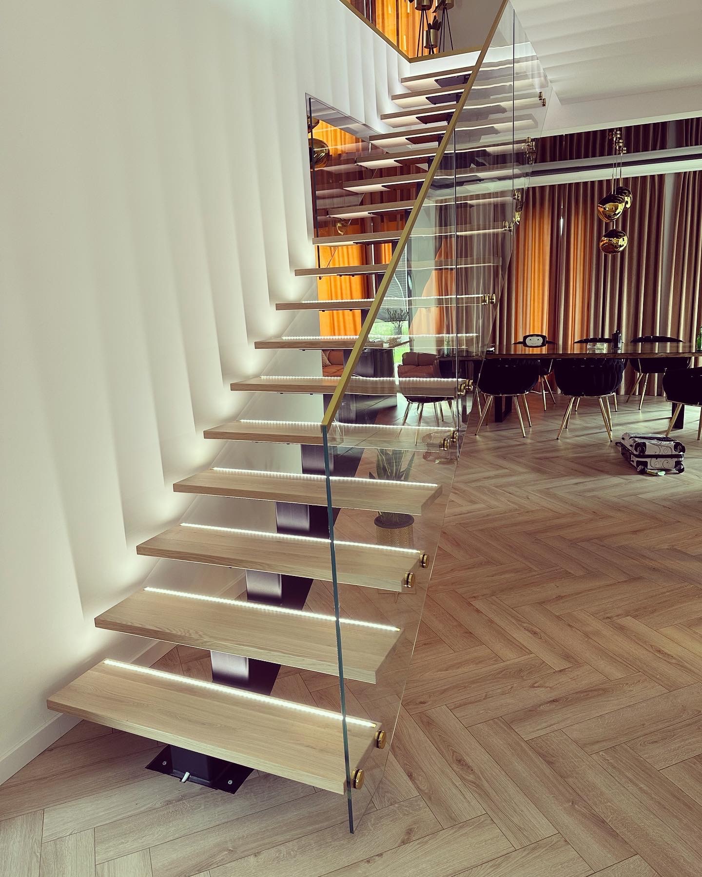 Nowoczesne schody z drewnianymi stopniami, podświetleniem LED i szklaną balustradą, z metalowymi elementami w kolorze złotym. W tle widoczny stół i krzesła.