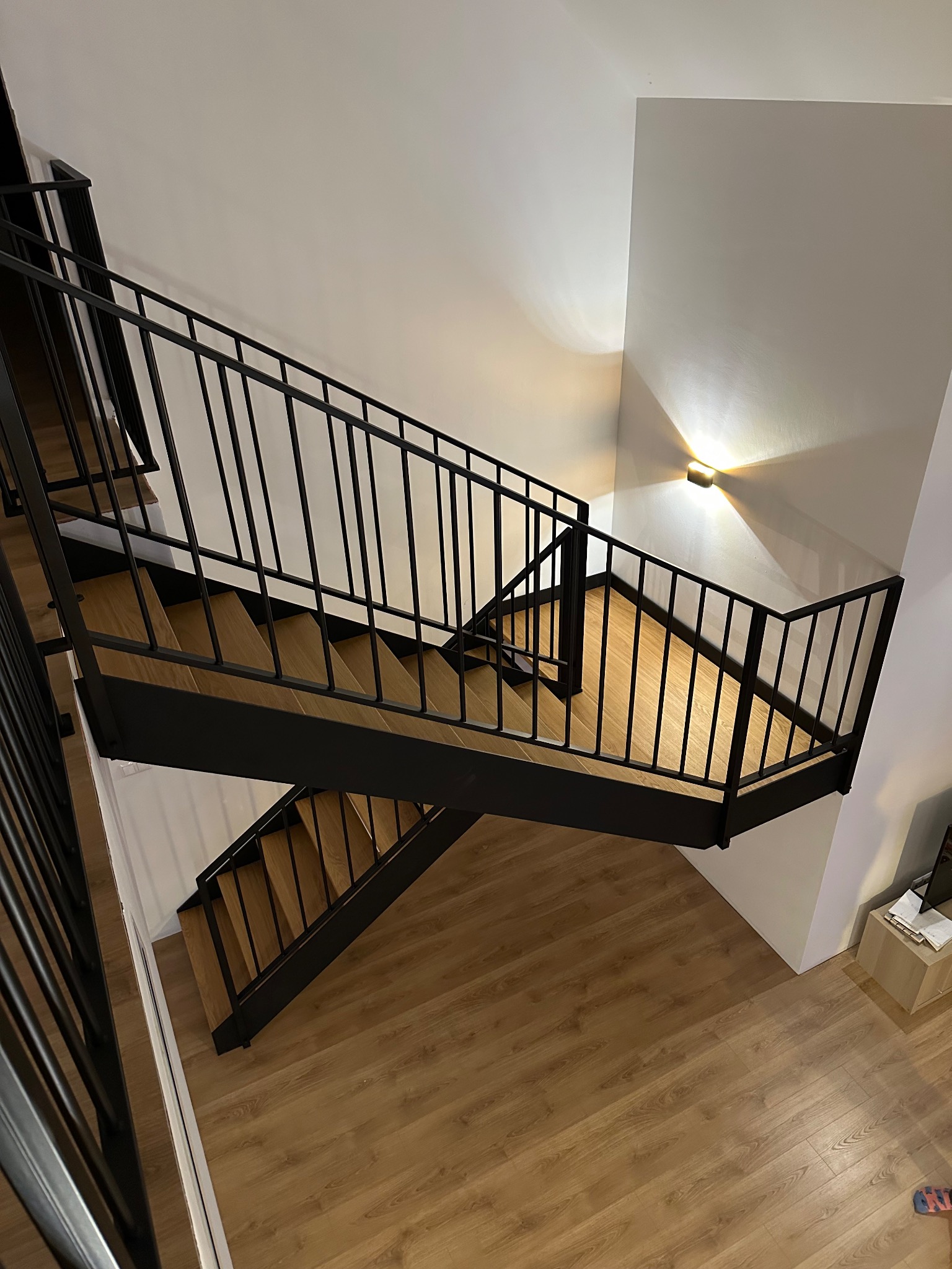 Nowoczesne schody z drewnianymi stopniami i czarną metalową balustradą, oświetlone punktowym światłem, widok z góry na drewnianą podłogę.