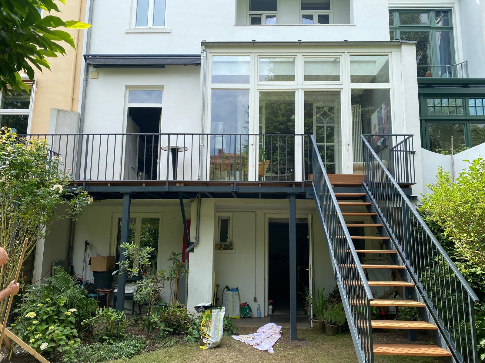 Zewnętrzne schody z metalową konstrukcją i drewnianymi stopniami prowadzą na balkon z metalową balustradą. Widoczne rośliny w ogrodzie i fragment budynku z oknami.