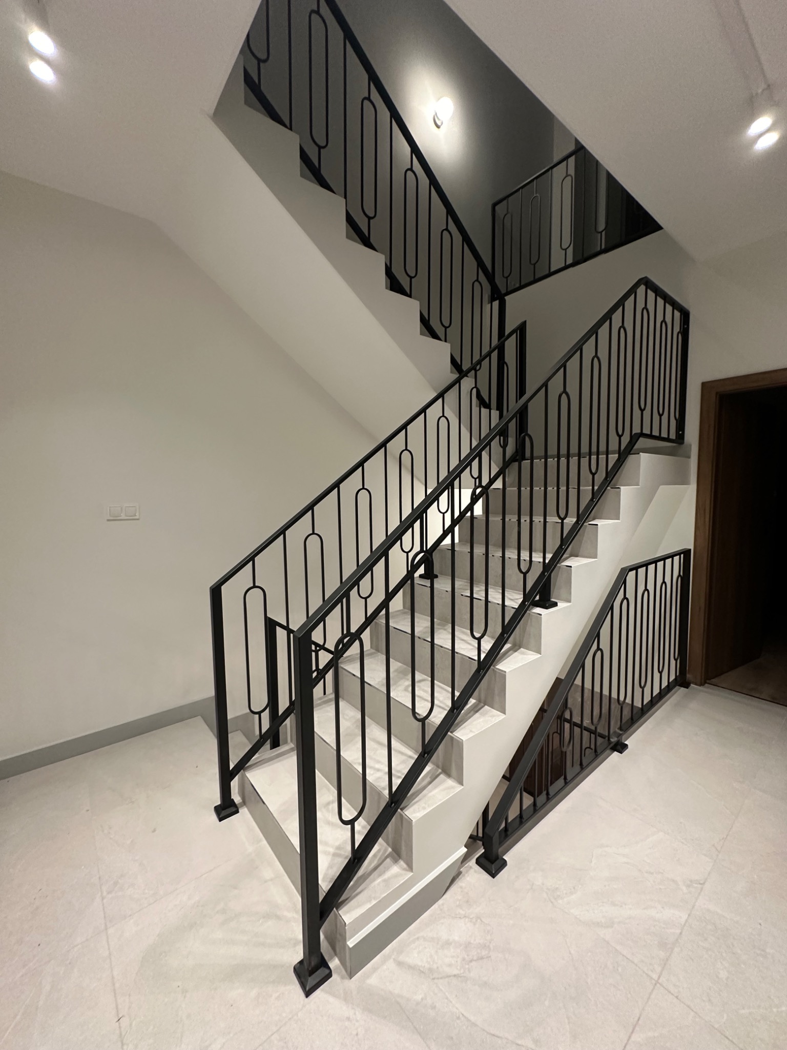 Wewnętrzne schody z czarną metalową balustradą o dekoracyjnych, zaokrąglonych elementach. Jasne płytki na stopniach i podłodze. Minimalistyczny design.