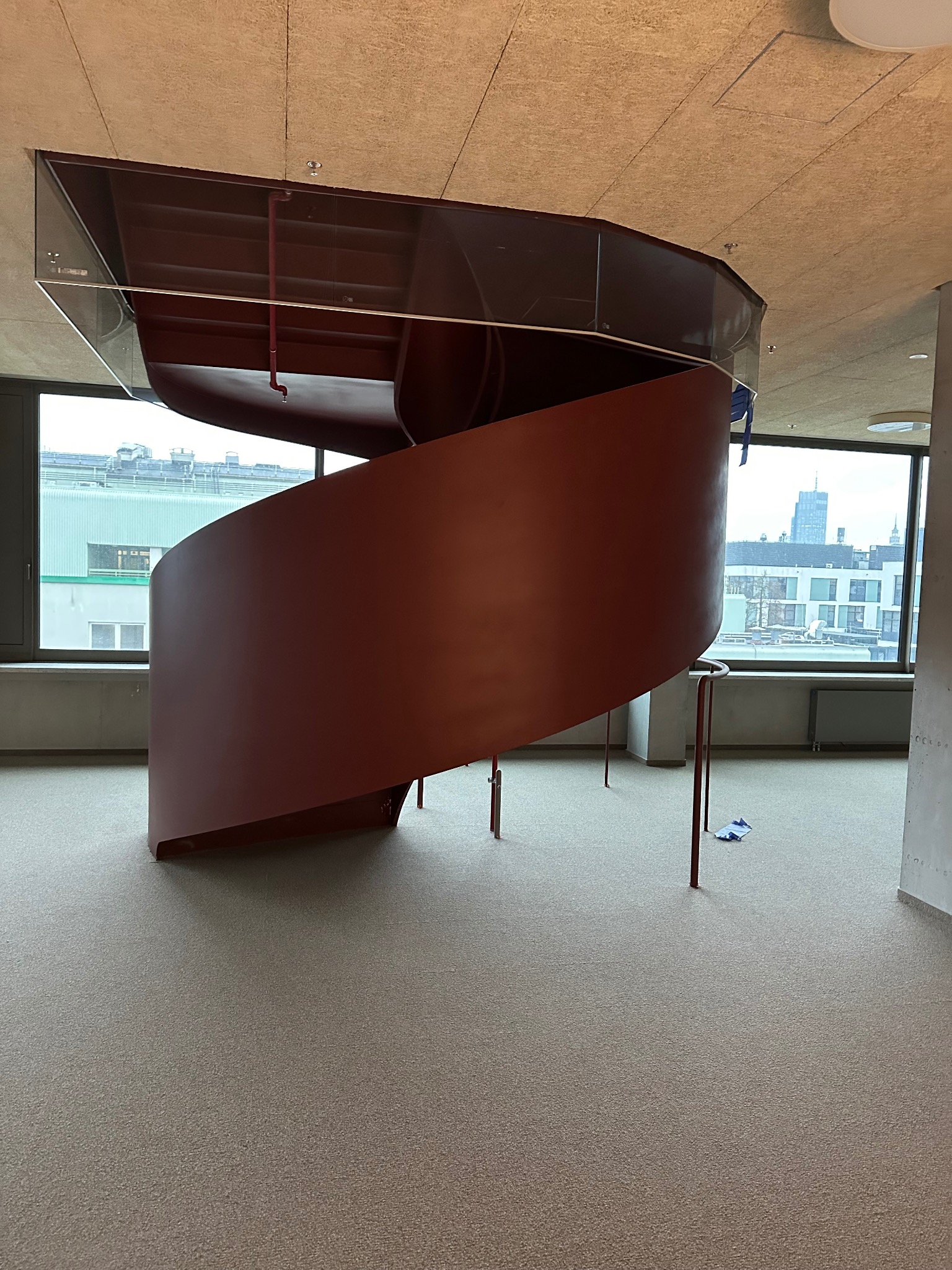 Spiralne schody metalowe w nowoczesnym wnętrzu, z widokiem na miasto za oknem. Minimalistyczny design, jasna podłoga, sufit z fakturą. Czerwona konstrukcja schodów.