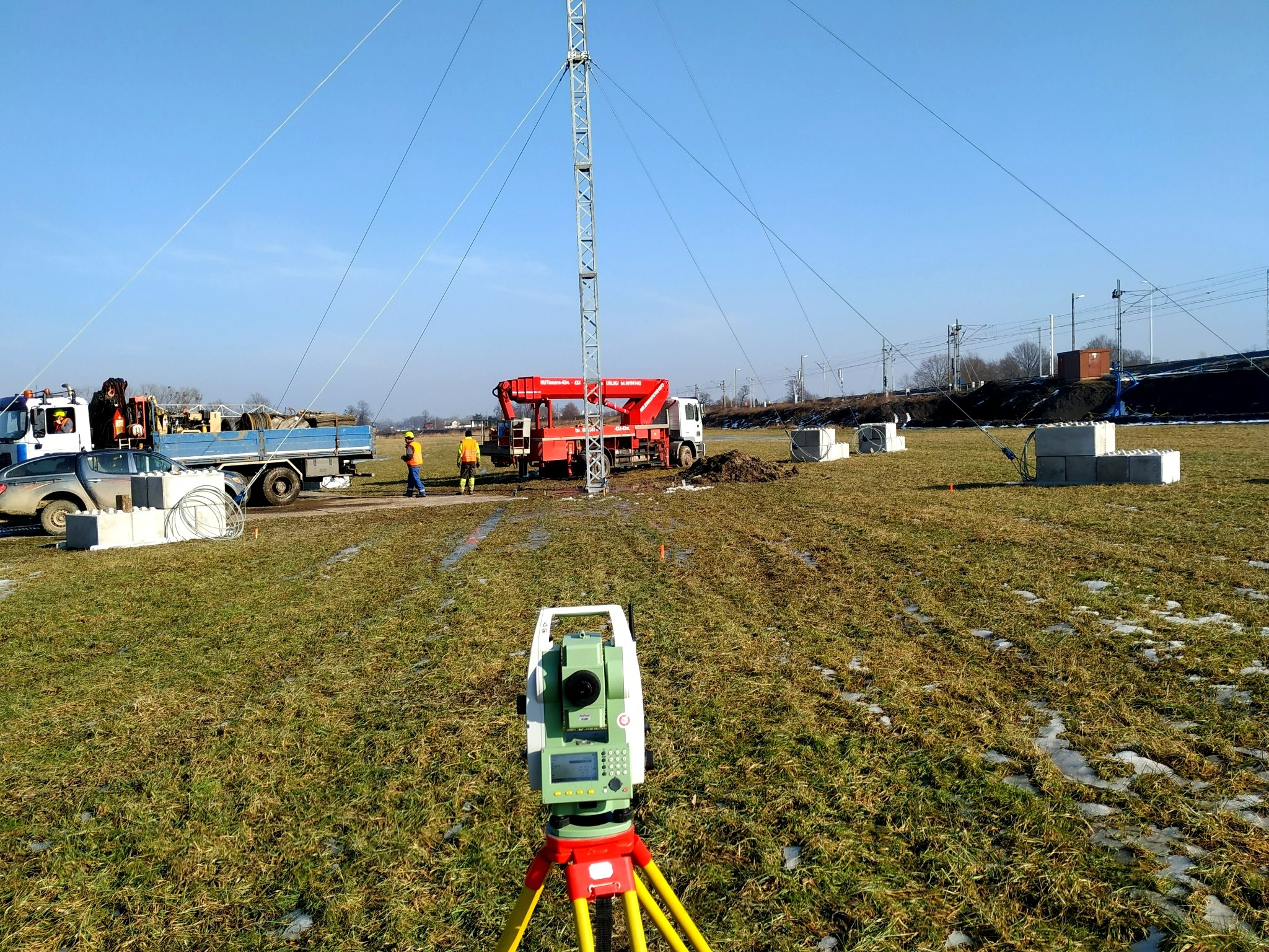 Pomiar geodezyjny z użyciem niwelatora na tle budowy masztu telekomunikacyjnego. Widoczny sprzęt pomiarowy, teren, pojazdy i pracownicy w oddali.