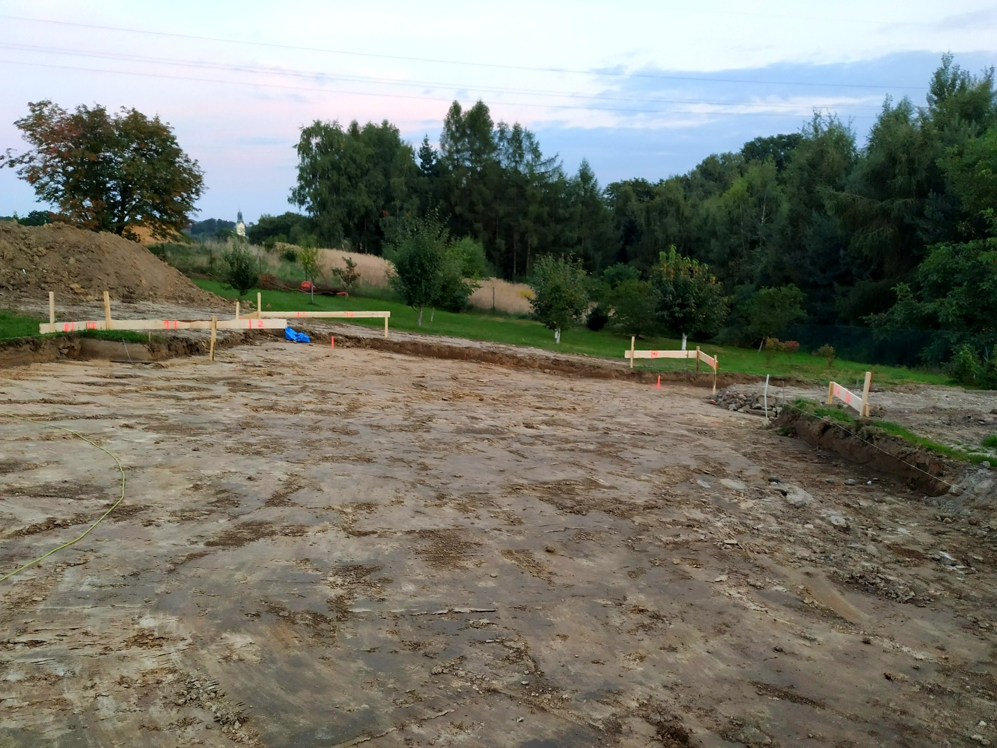 Wytyczony teren pod budowę z drewnianymi palikami i sznurkiem, w tle drzewa i pagórek. Widoczne ślady po pracy maszyn.