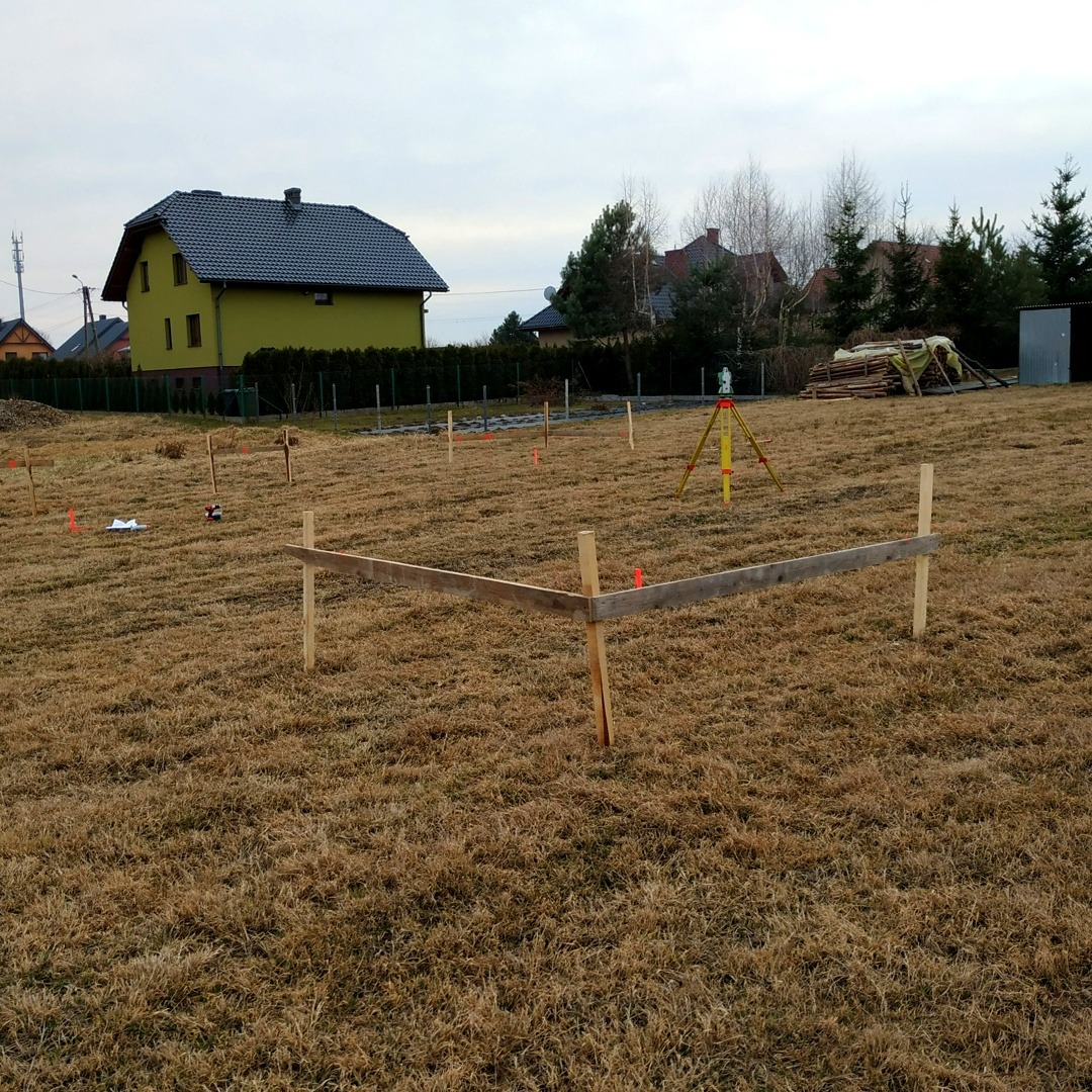 Tyczenie działki w Połomi: drewniane słupki wyznaczają granice. W tle żółty niwelator i dom. Sucha trawa i pochmurne niebo.
