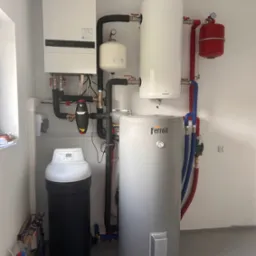 Kompletna instalacja grzewcza w kotłowni: biały piec, zbiorniki Galmet i Ferroli, czarny filtr, rury w izolacji. Widoczne zawory i manometry.