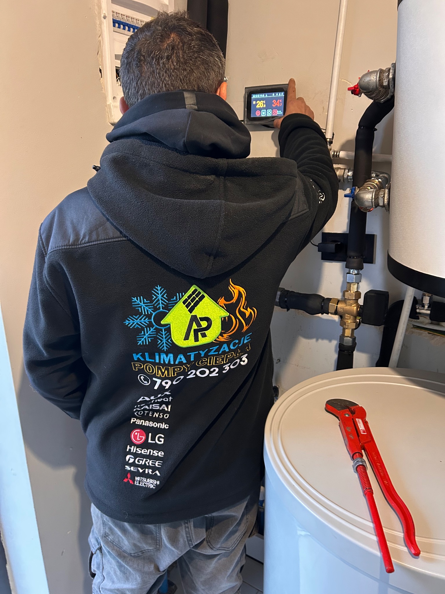 Instalator w bluzie firmowej reguluje panel pompy ciepła. Widoczne rury, zawory i narzędzie. Specyficzne logo firmy: klimatyzacja i pompy ciepła.