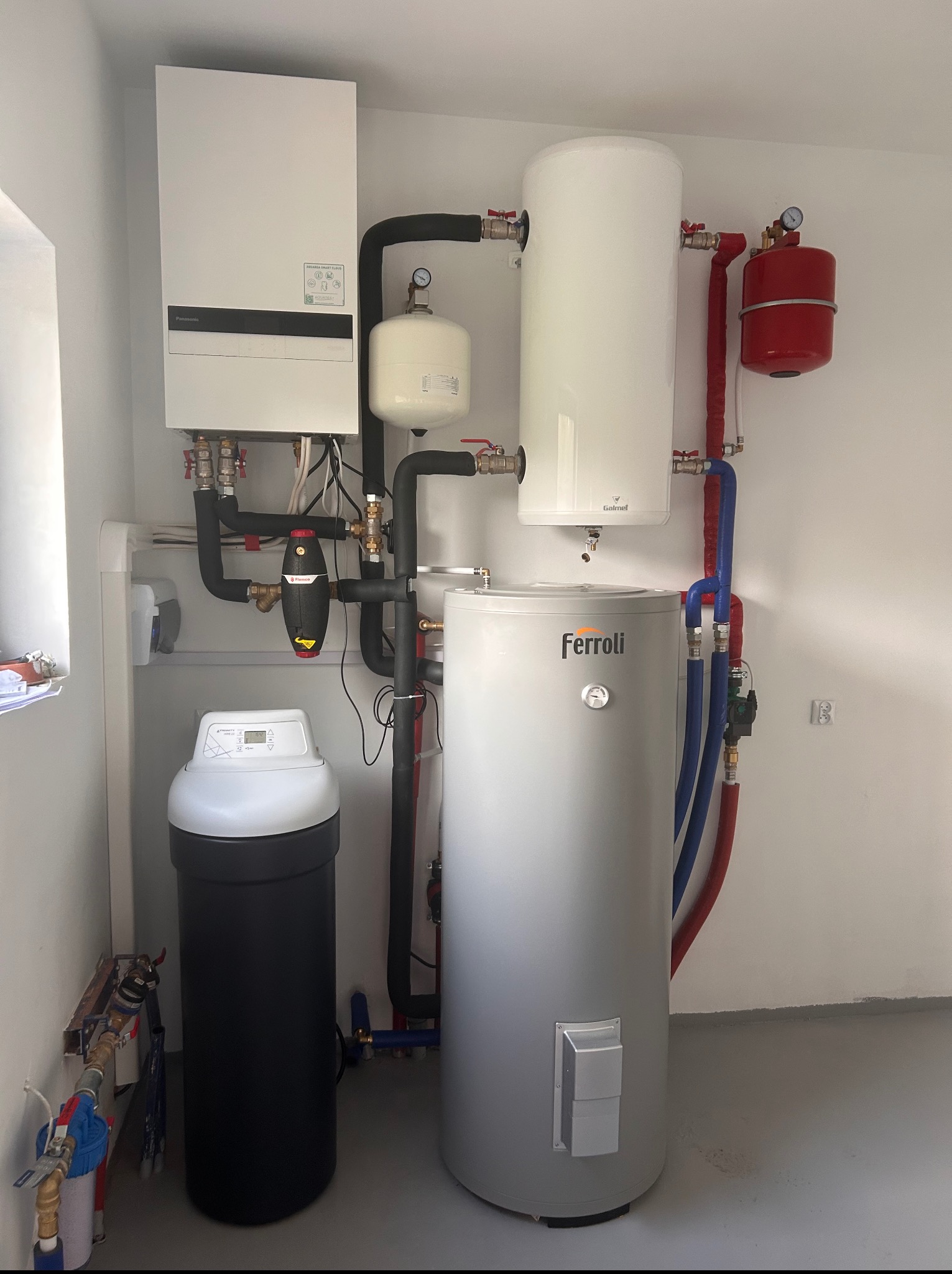 Kompletna instalacja grzewcza w kotłowni: biały piec, zbiorniki Galmet i Ferroli, czarny filtr, rury w izolacji. Widoczne zawory i manometry.