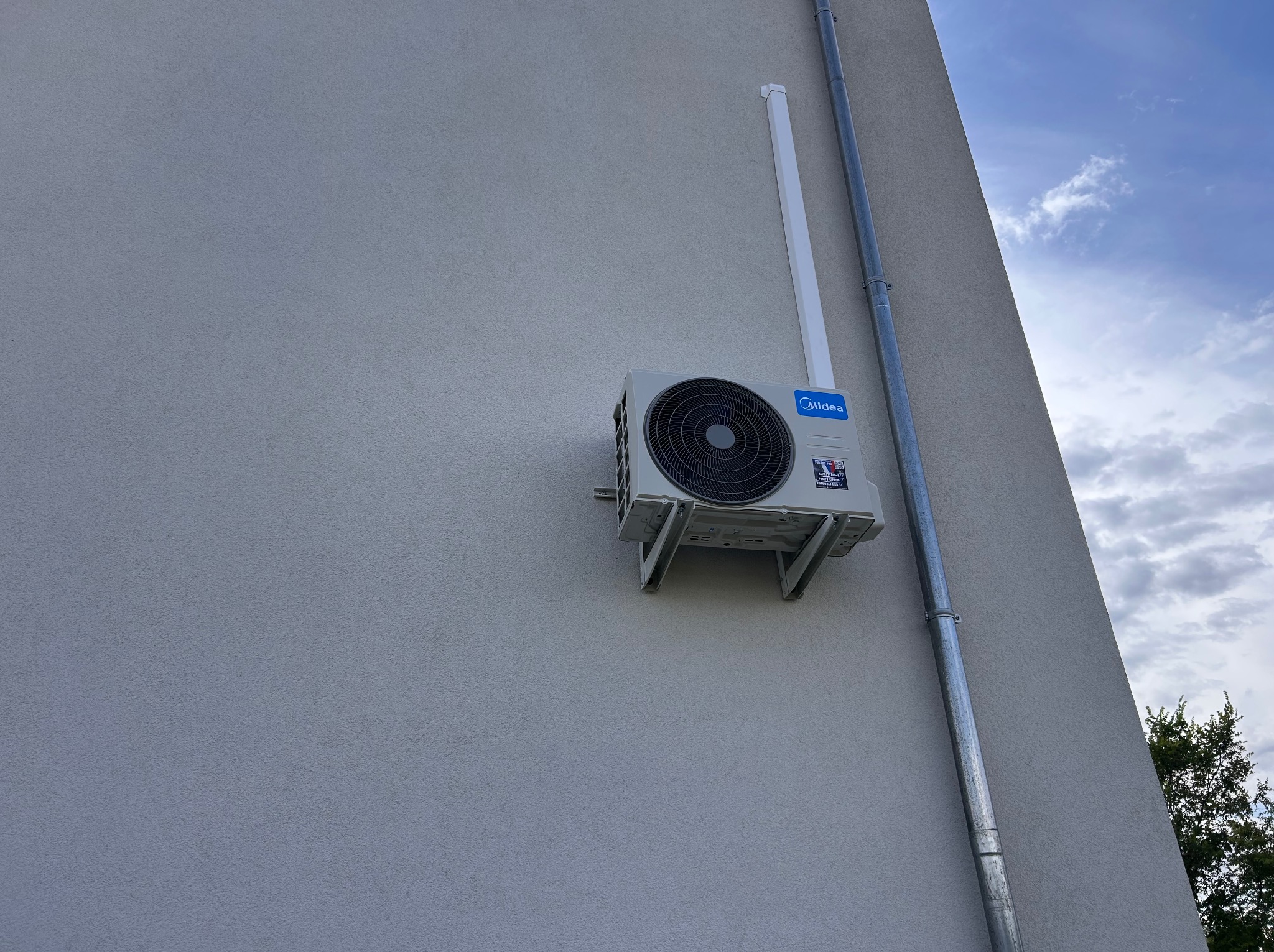 Zamontowana klimatyzacja Midea na szarej elewacji budynku, widoczna rura odprowadzająca skropliny i fragment nieba. Ujęcie z dołu.