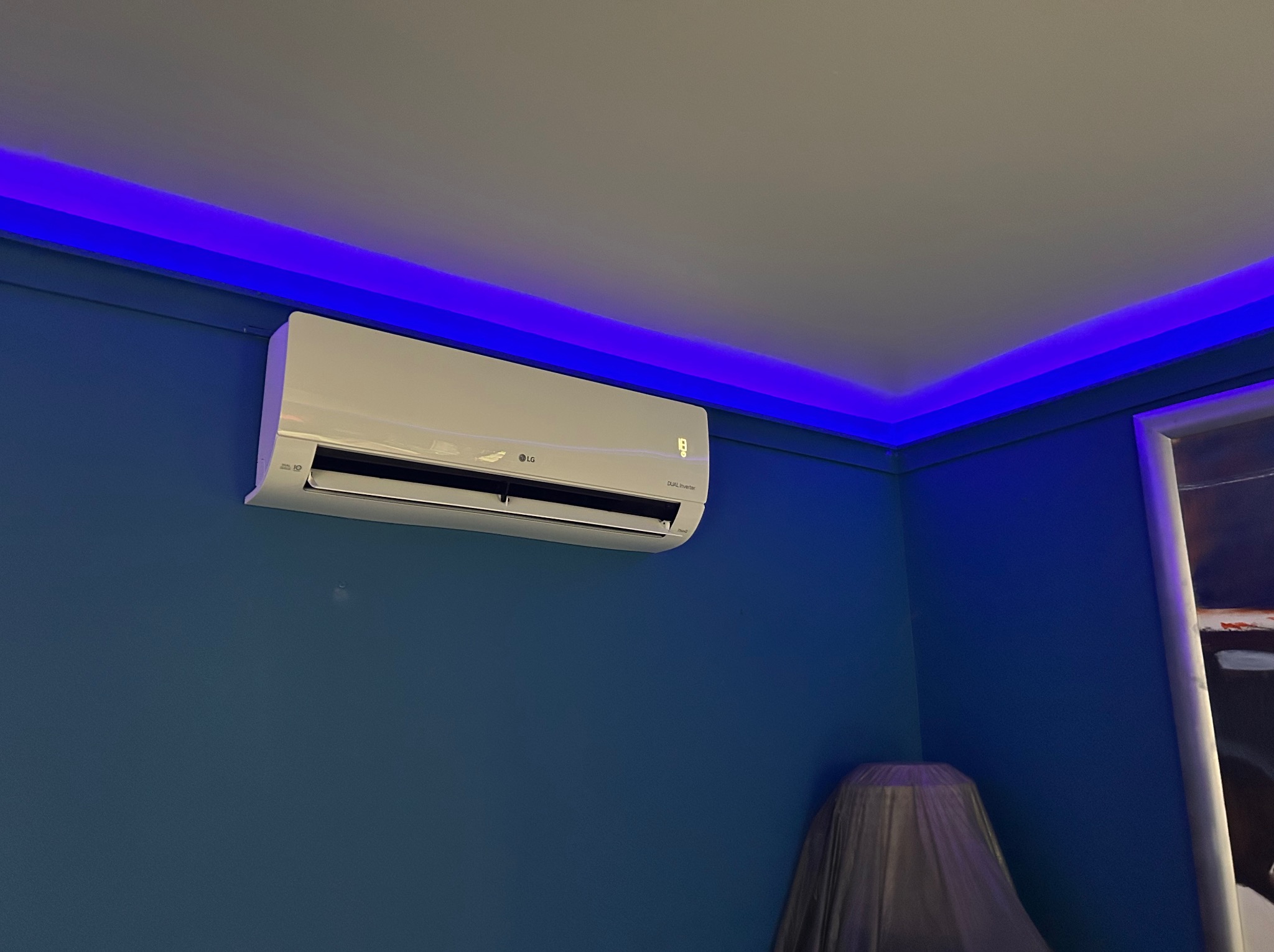 Biała klimatyzacja LG na niebieskiej ścianie, oświetlona niebieskim światłem LED na suficie. Widoczny fragment okna i mebla pod folią ochronną.