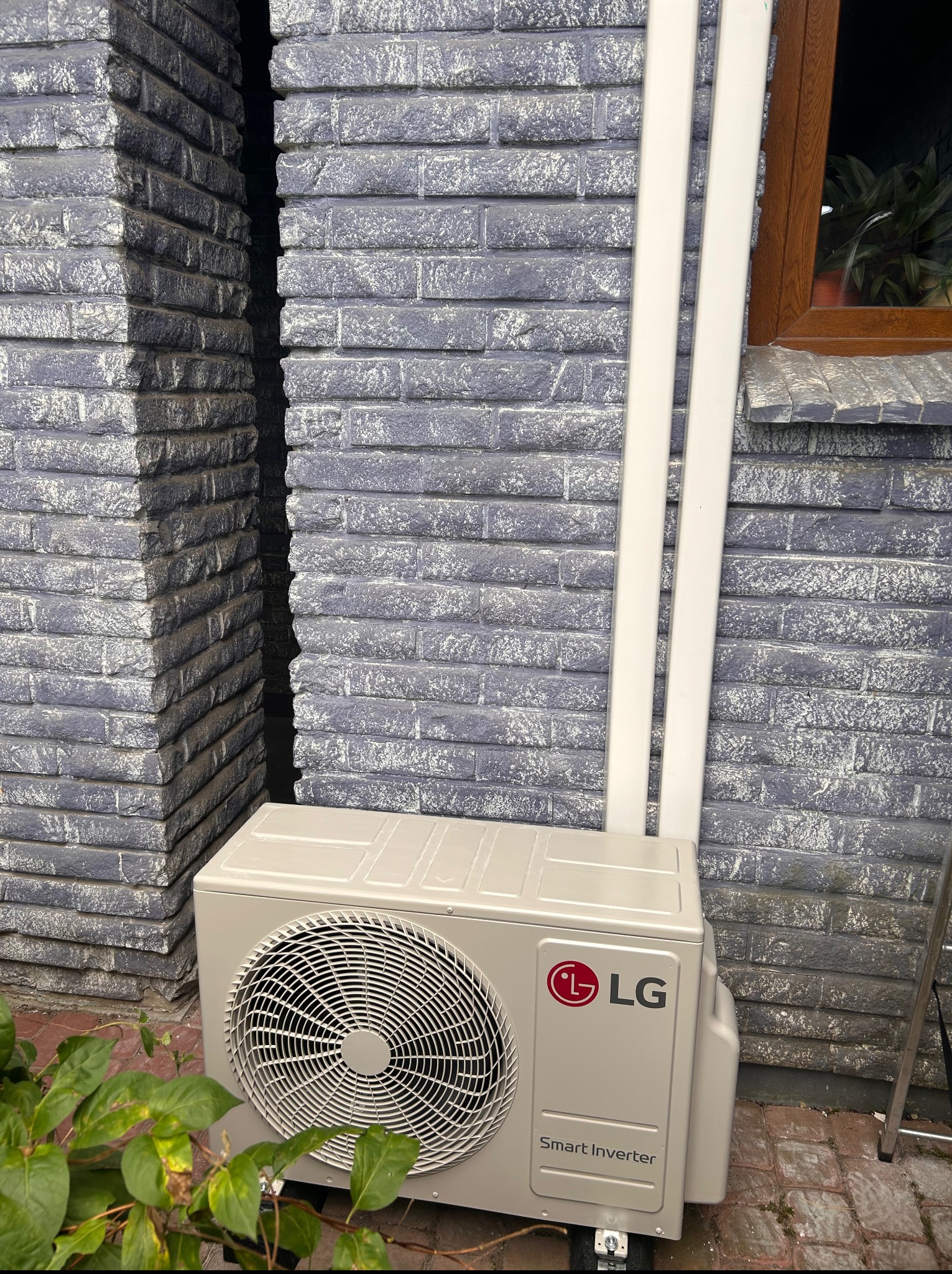 Zamontowana jednostka zewnętrzna klimatyzacji LG Smart Inverter na tle szarej, ceglanej ściany budynku z roślinnością u podstawy. Widoczne białe rury odprowadzające.