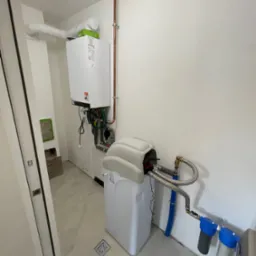 Biały piec gazowy z miedzianą instalacją i filtrem wody w jasnym pomieszczeniu. Widoczne rury, zawory i elementy przyłączeniowe. Minimalistyczny design.