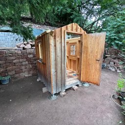 Usługi ogrodnicze Andrii Sova - Drewniana altana z otwartymi drzwiami w ogrodzie, otoczona kamiennym murem i zielenią. Widoczne detale konstrukcyjne i naturalna faktura drewna.