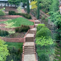 Usługi ogrodnicze Andrii Sova - Kaskadowy ogród z trawnikiem, schodami z kostki i ceglanymi murkami. W tle widoczni pracownicy podczas prac ziemnych.