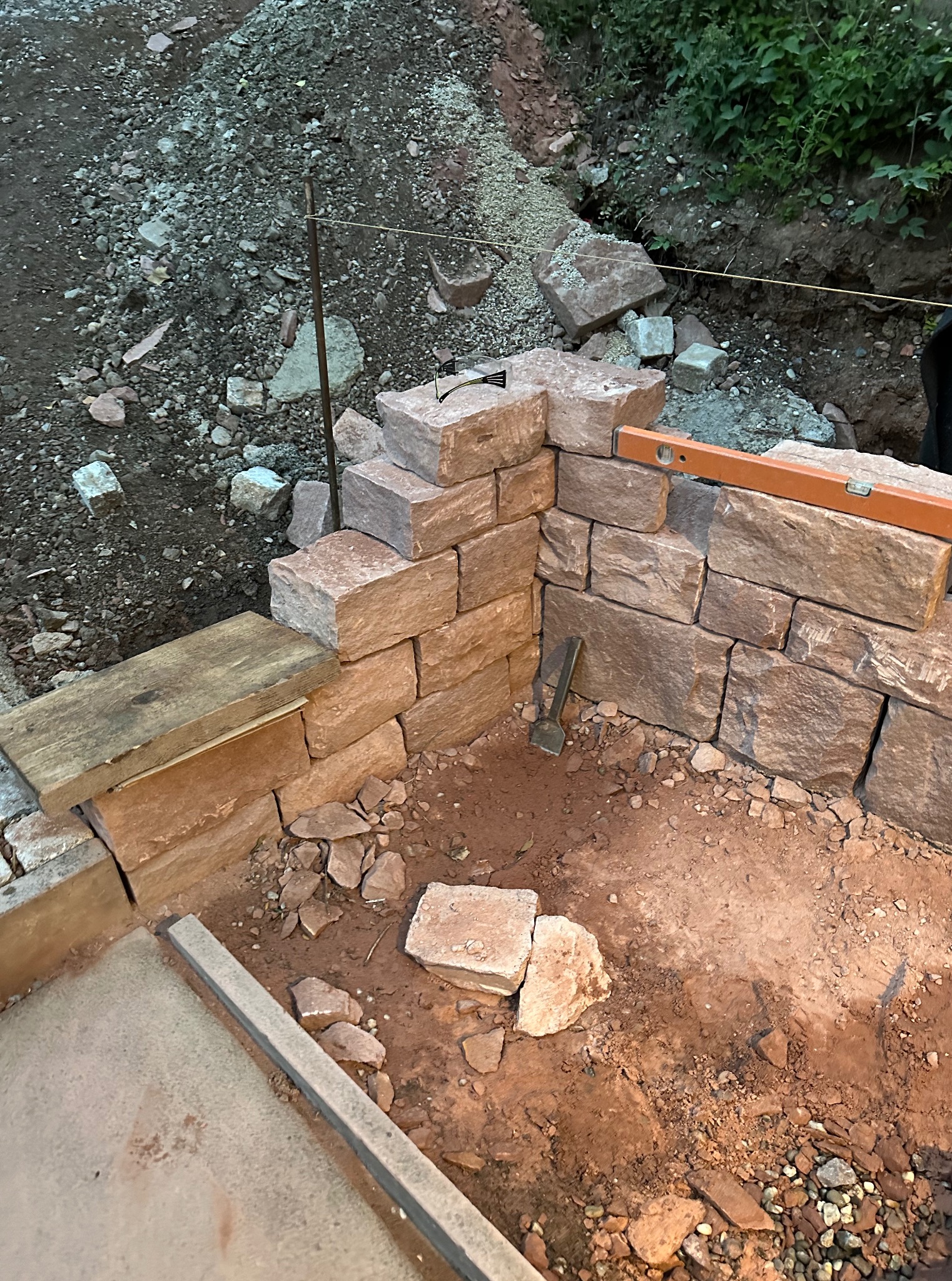 Budowa muru oporowego z kamienia: fragment konstrukcji z poziomnicą i narzędziem, widoczne materiały i etap prac ziemnych w ogrodzie.