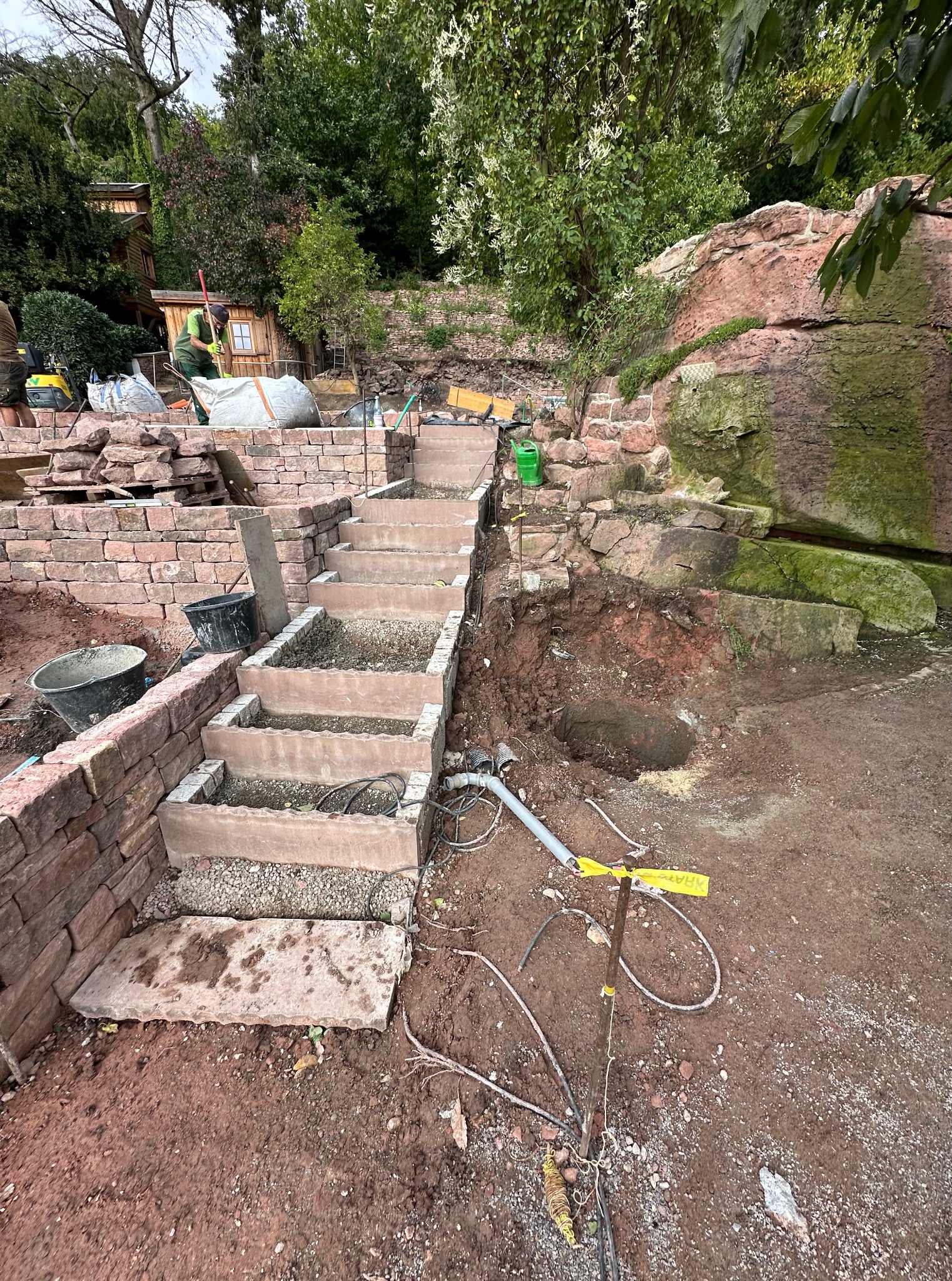 Budowa kamiennych schodów ogrodowych z wypełnieniem żwirowym, w tle zielona roślinność i elementy krajobrazu. Widoczne narzędzia i materiały budowlane.