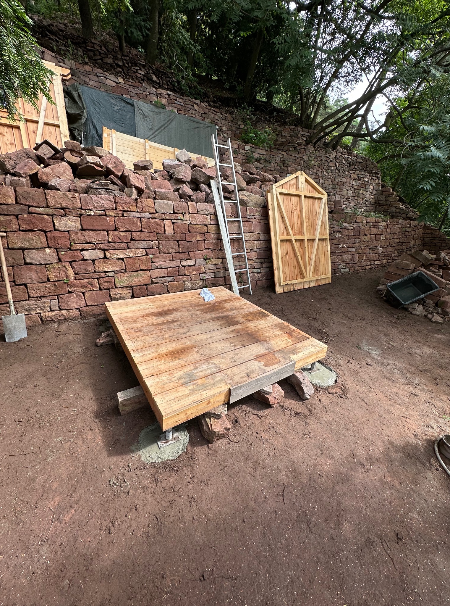 Budowa tarasu na nierównym terenie: drewniana platforma na betonowych podporach, kamienny murek w tle, drabina i elementy drewniane. Widoczne narzędzia i etap prac.